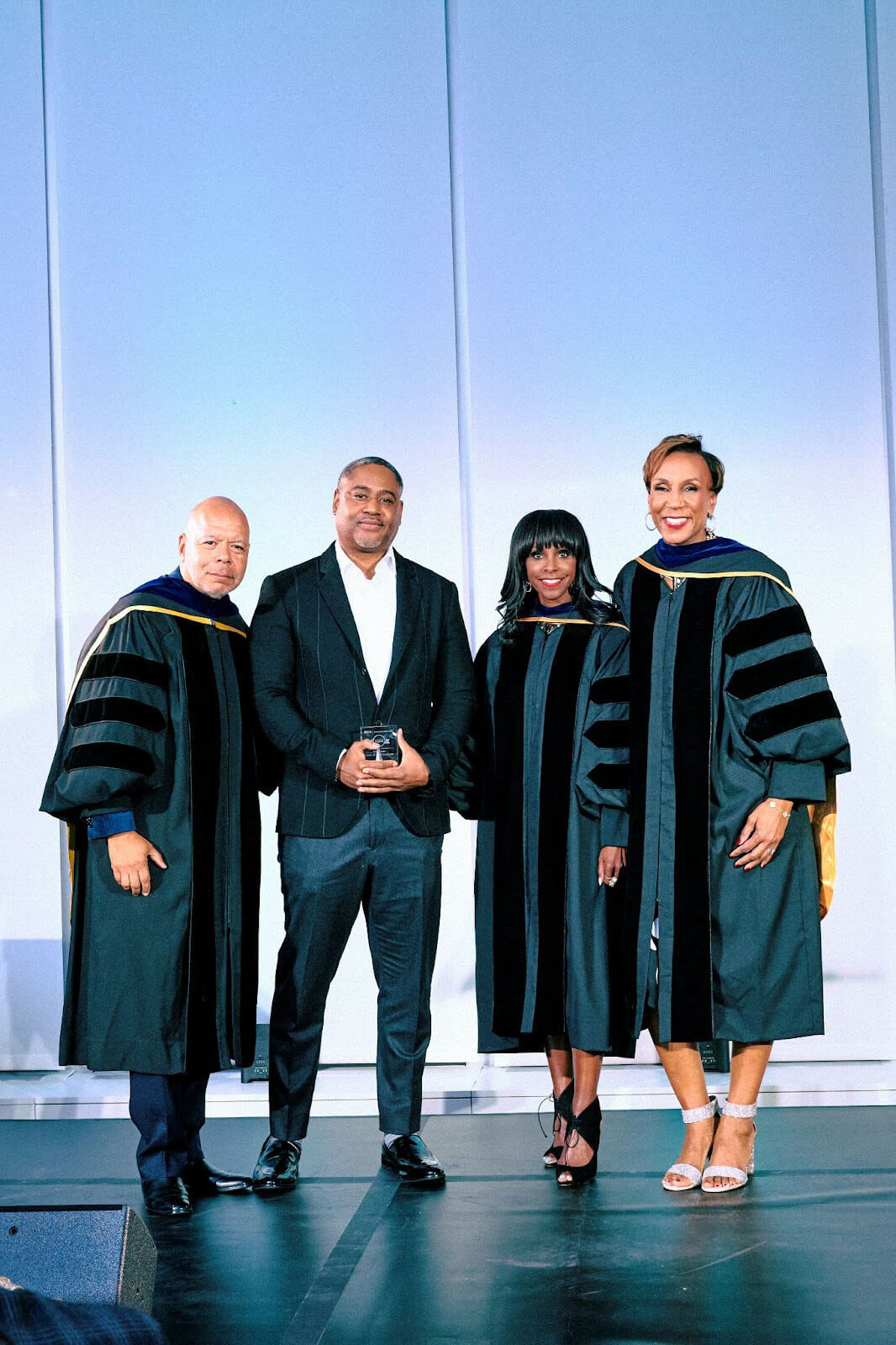 Four individuals, three in academic regalia and one in a black suit, posing for a photo on stage with an award.