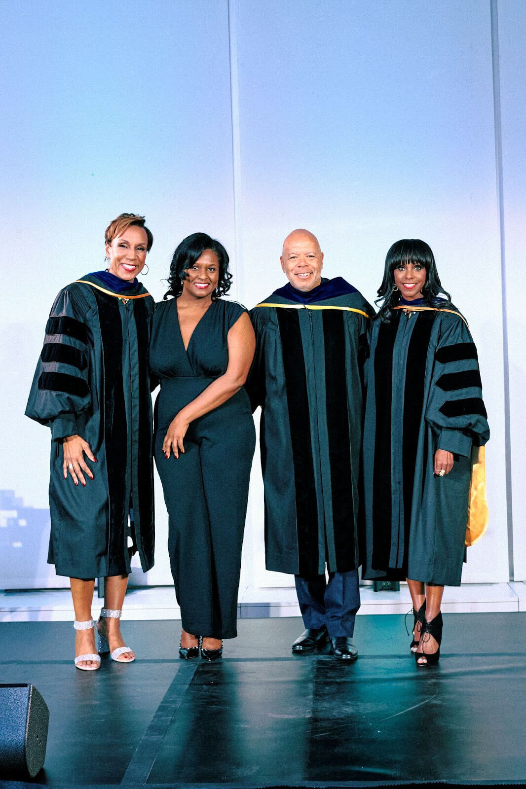 Four people standing together on stage, three in academic robes and one in a black dress, smiling for the photo.