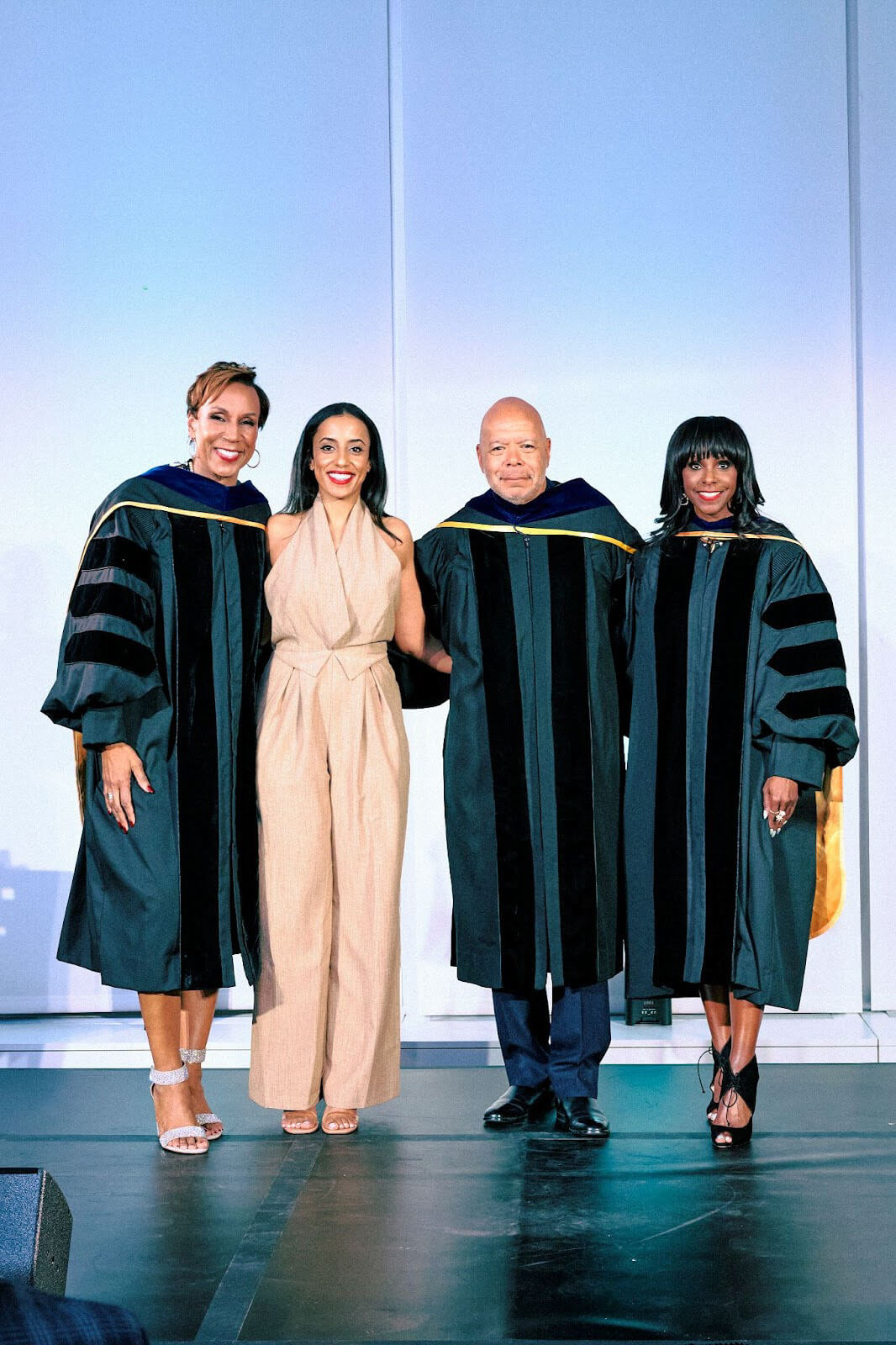 A group of four people on stage, three in academic robes and one in a tan jumpsuit, smiling and posing.