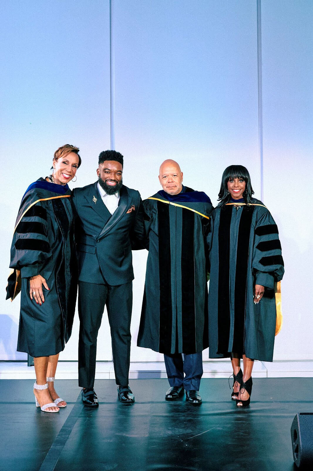 A group of four people, three in academic robes and one in a dark suit with a bow tie, smiling on stage.