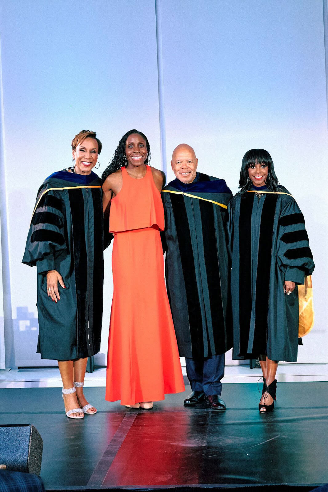 Four people posing for a group photo on stage, with one in a bright orange dress and three in academic regalia.