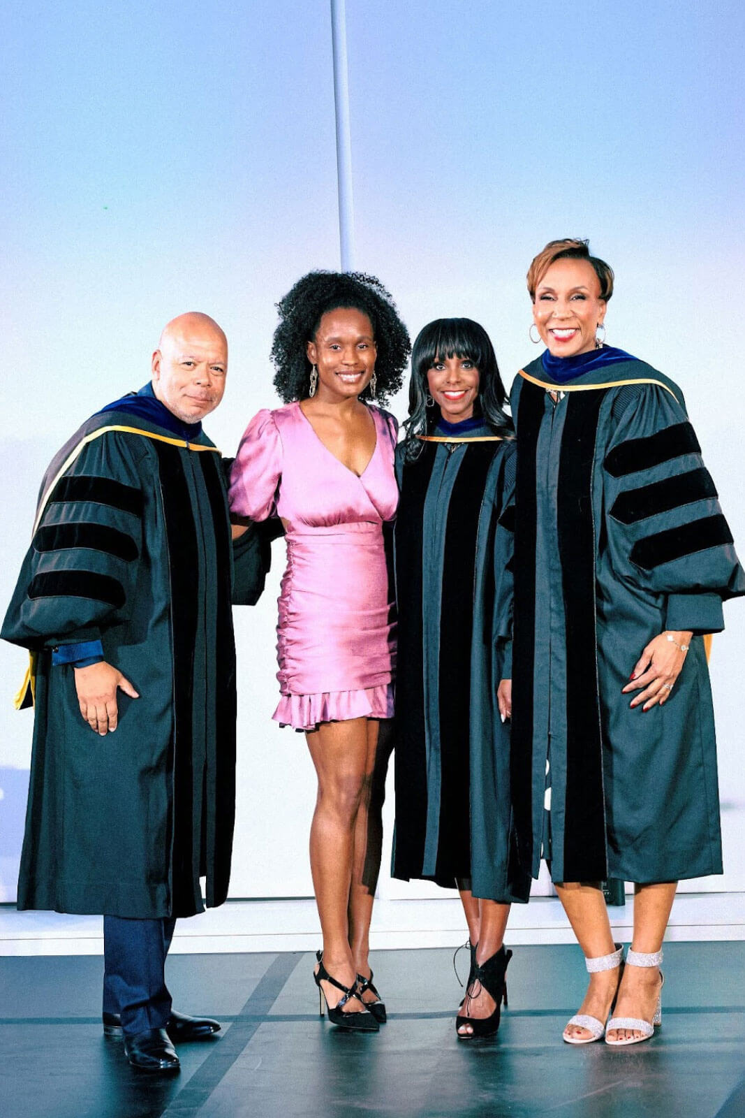 A group photo featuring three individuals in academic robes and one in a pink dress, smiling on stage.
