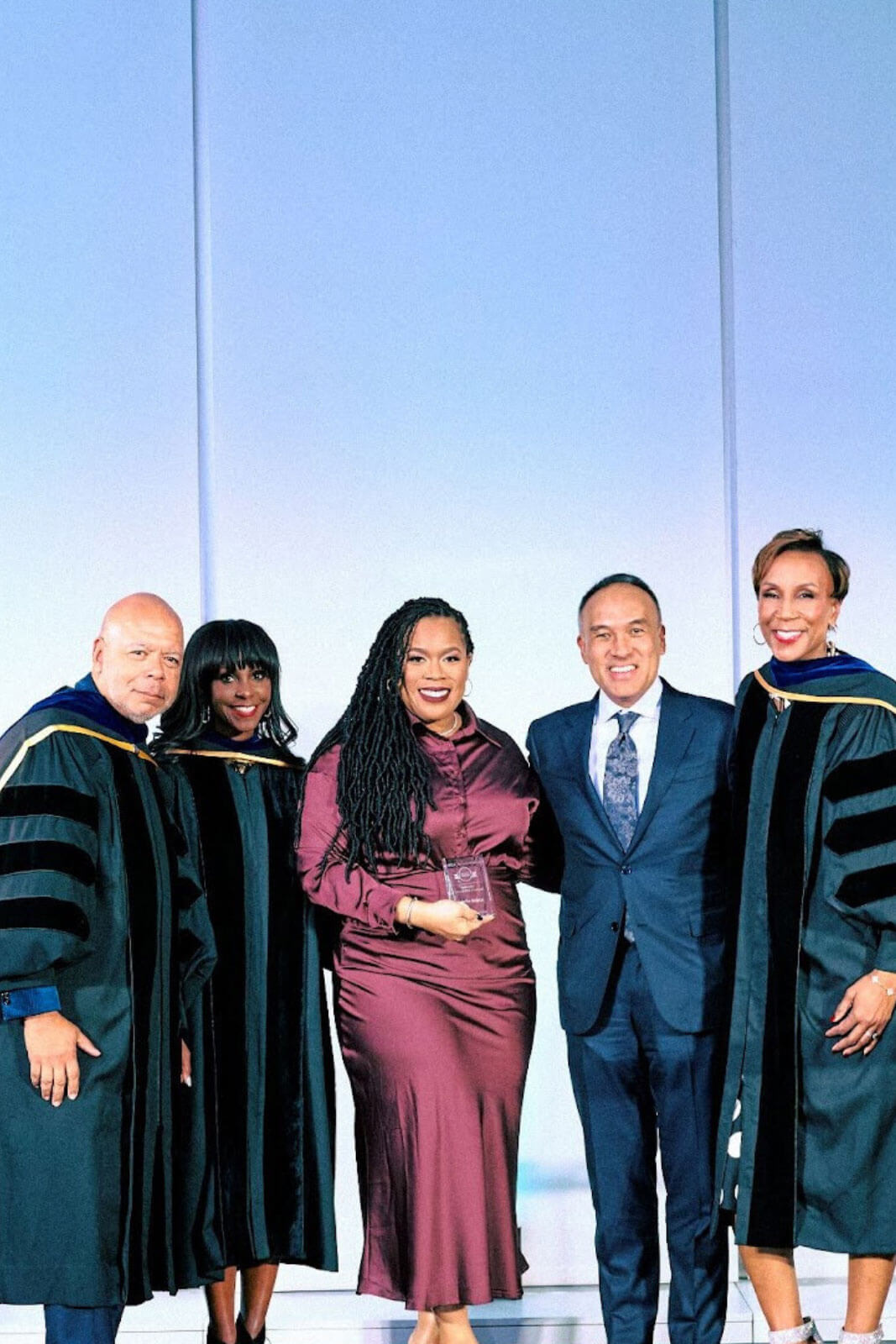 Five people on stage, one in a burgundy dress holding an award, accompanied by three in academic robes and one in a suit.