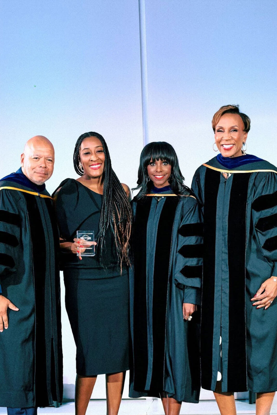 A group of four individuals, two in academic robes, standing together on a stage smiling, with one holding a glass award.