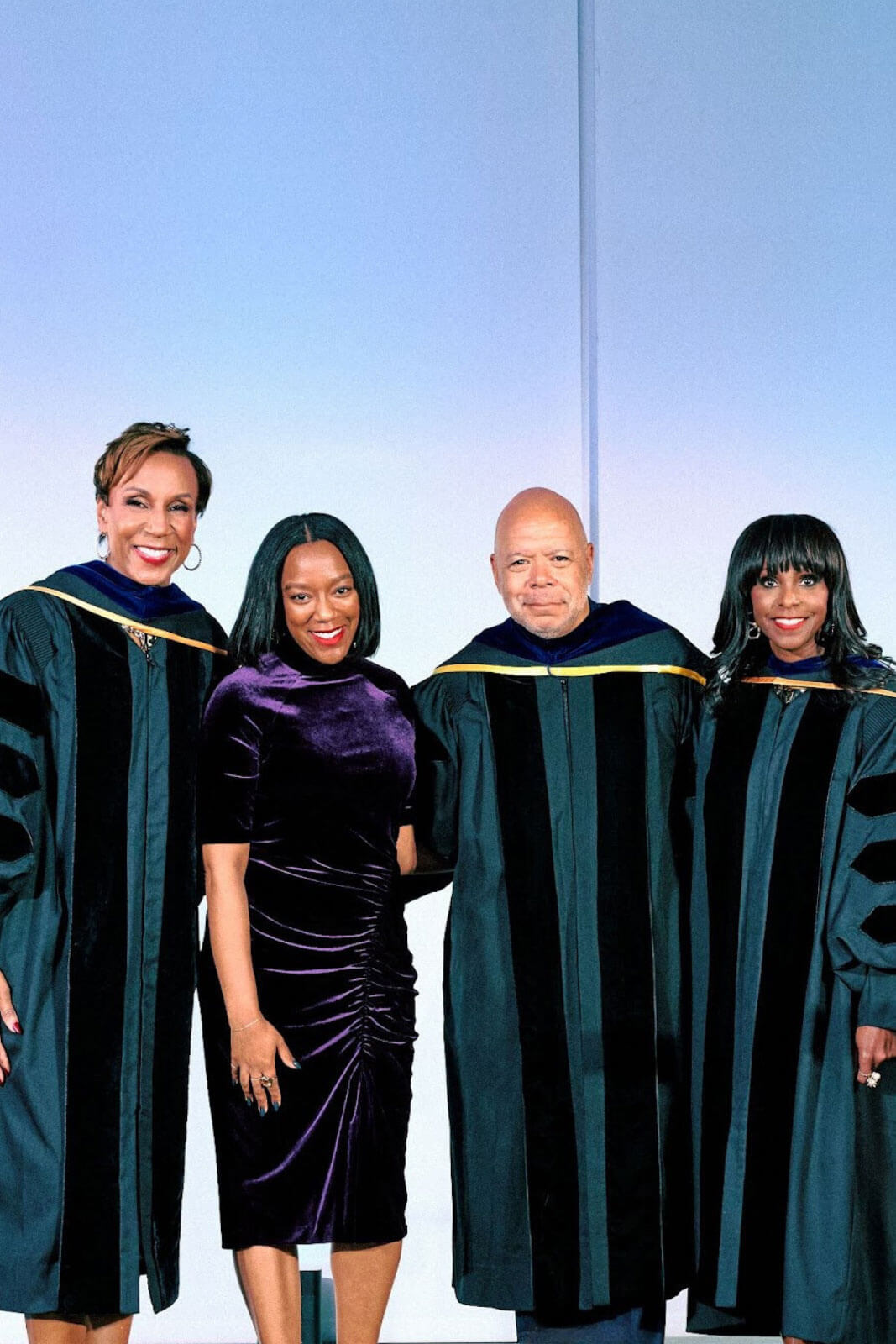 Another group of four, including two in academic robes and one in a deep purple velvet dress, posing with smiles and holding an award on a stage.