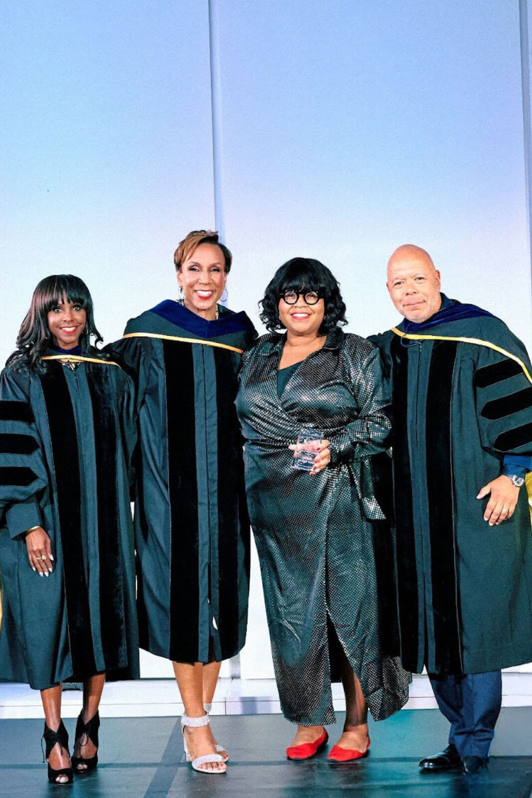 Four people, including two in academic robes, standing with a woman dressed in metallic attire holding a trophy on stage.