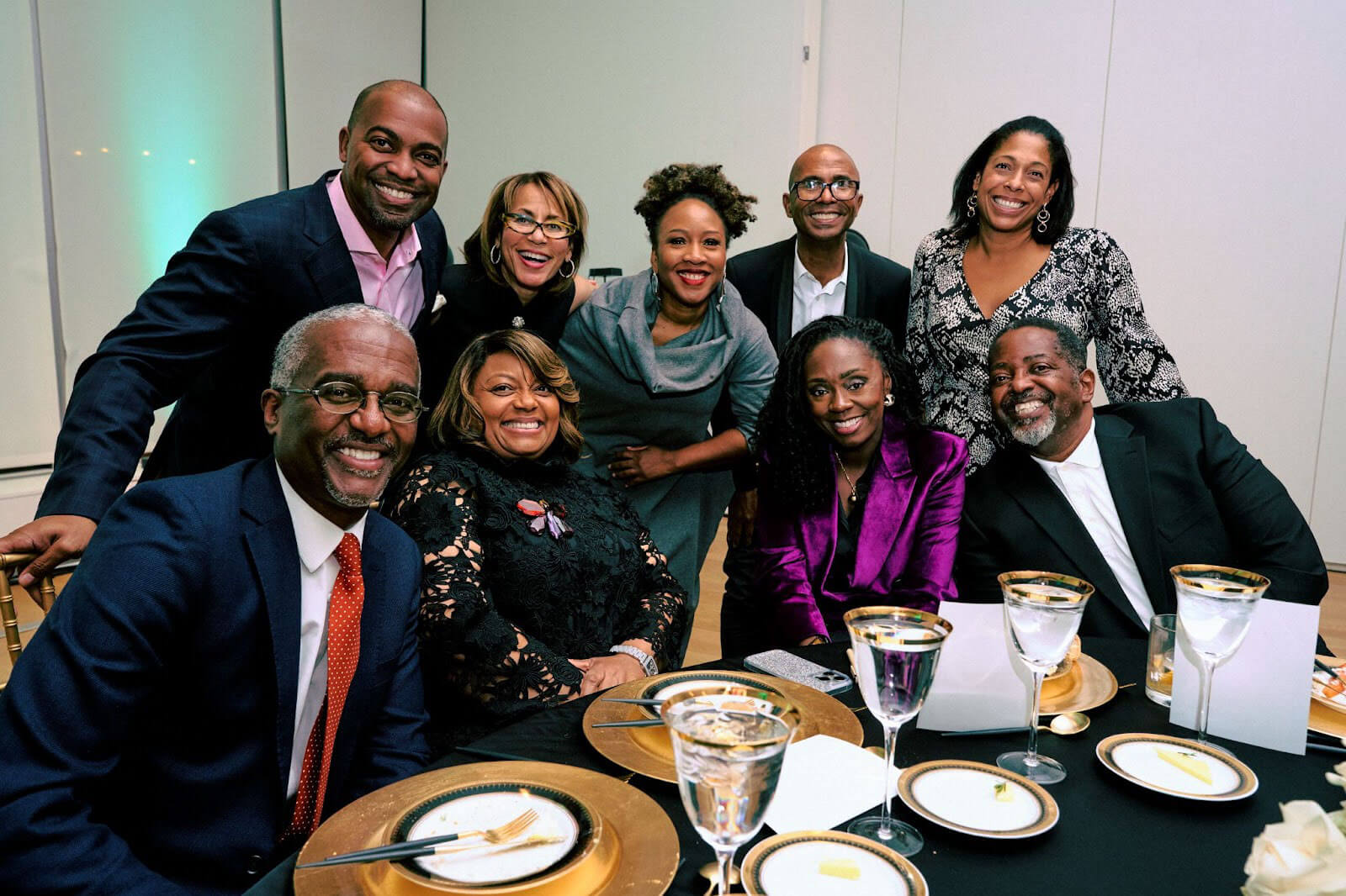 A group of individuals gathered around a table, smiling and enjoying a formal dining experience with gold-trimmed table settings.