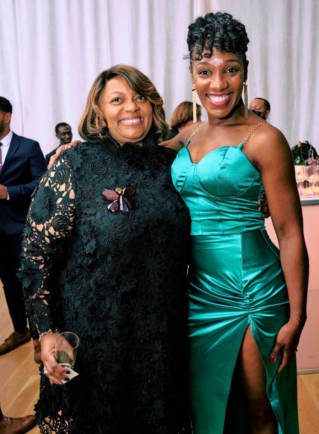 Two women smiling and posing together; one wearing a black lace dress and the other in an emerald green gown with a high slit.