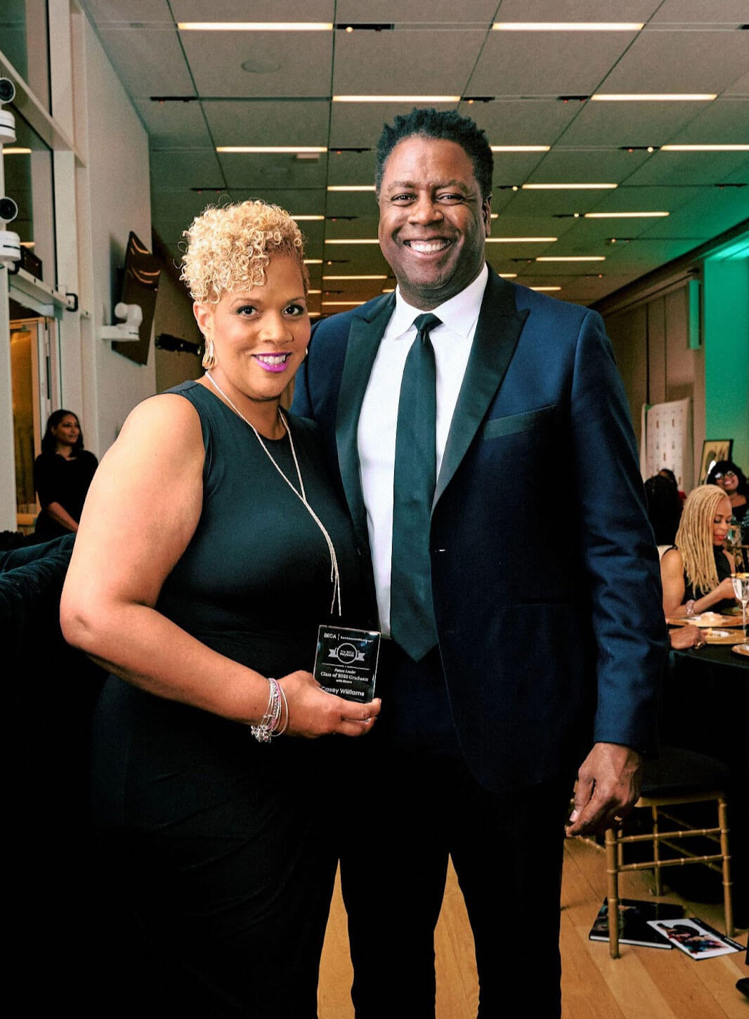 A man and a woman dressed in formal attire, posing together; the woman is holding an award.