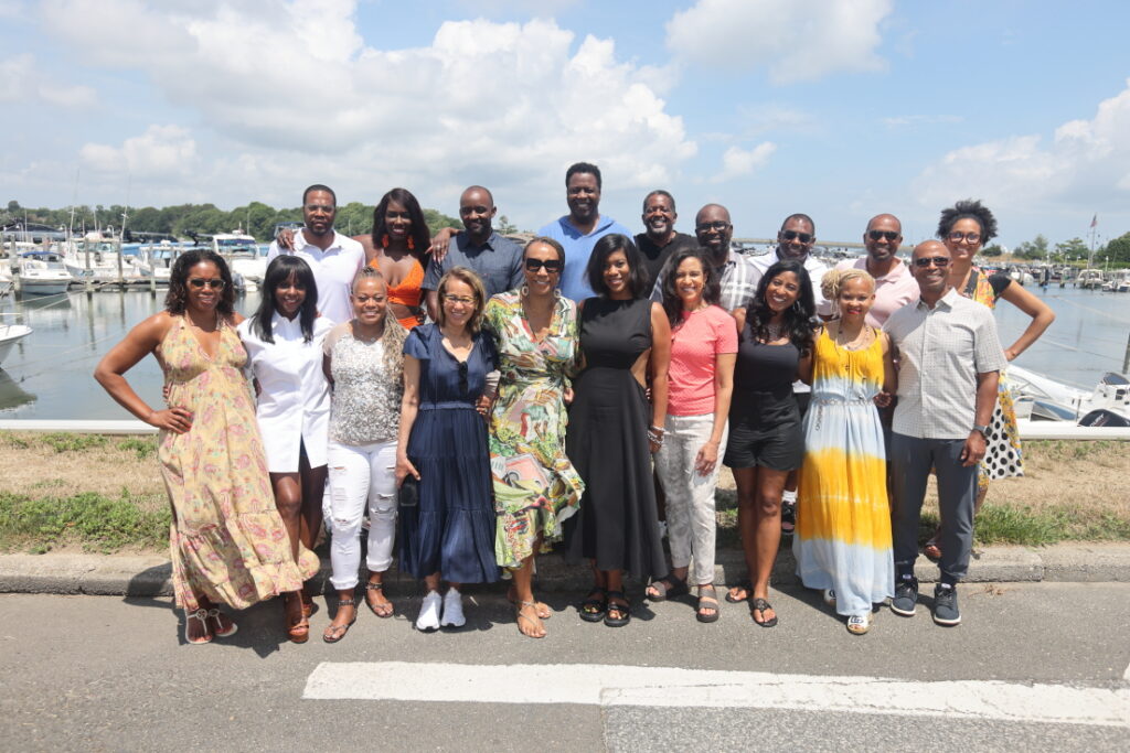 A large group of adults poses outdoors on a sunny day, standing in front of a marina with docked boats. The men and women wear casual summer clothing and smile at the camera.