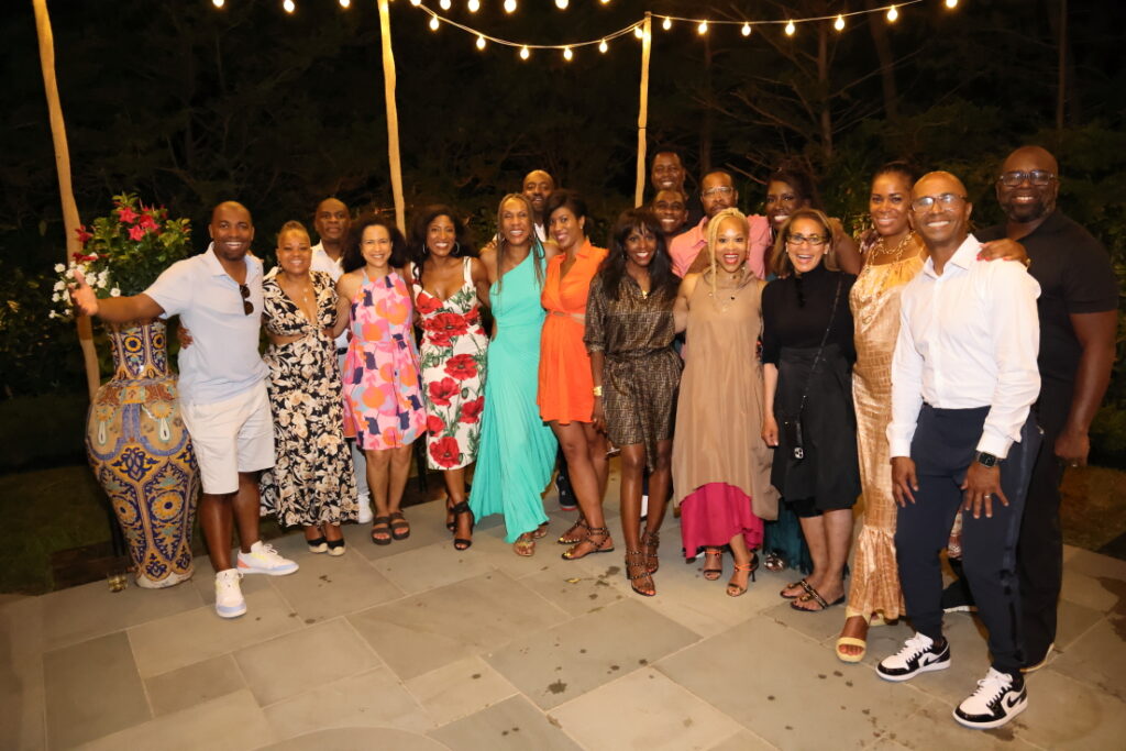 A large group of people poses under string lights at an outdoor nighttime gathering, all dressed in bright, summery outfits.