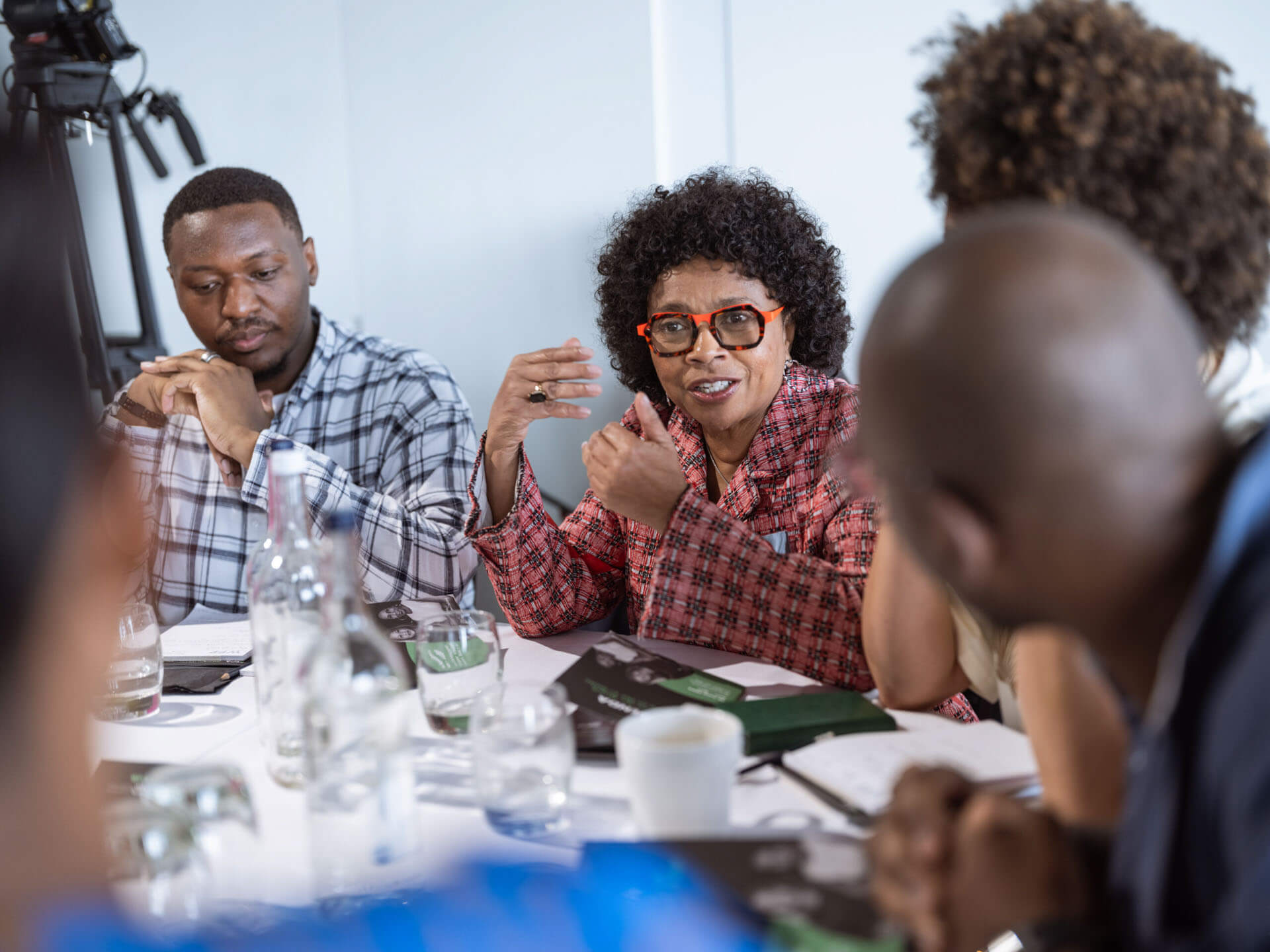 A woman in a patterned jacket and red glasses engages in a lively discussion at a table surrounded by others.