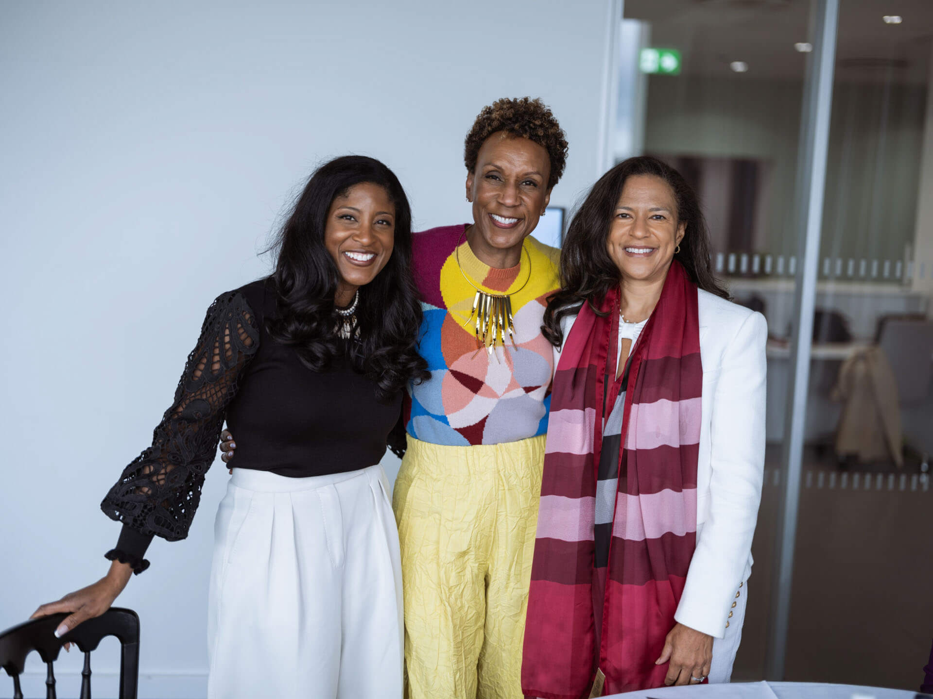 Three women, dressed stylishly, pose together with bright smiles in a conference setting.