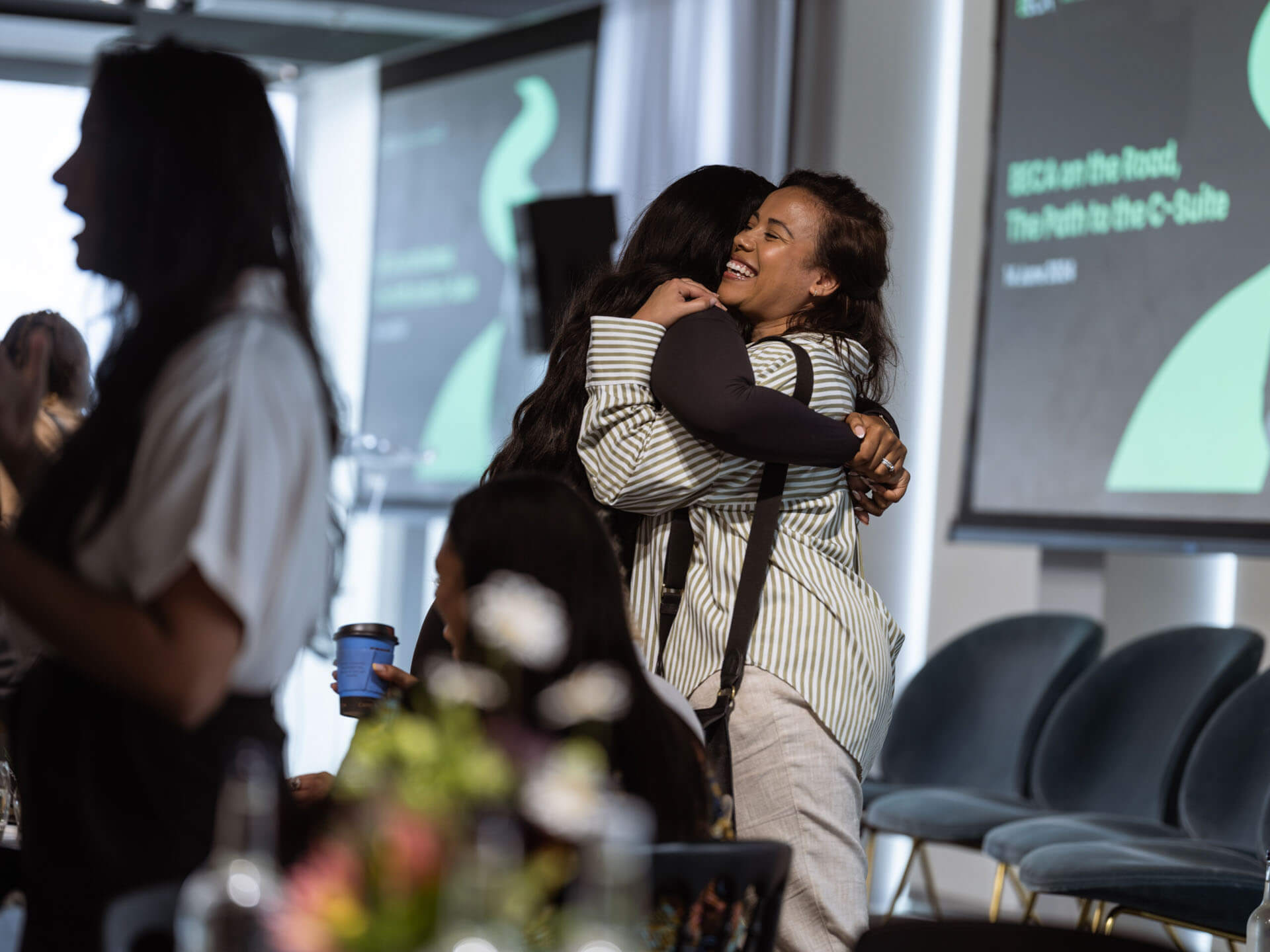 Two women sharing a heartfelt hug in a conference room with event signage in the background.