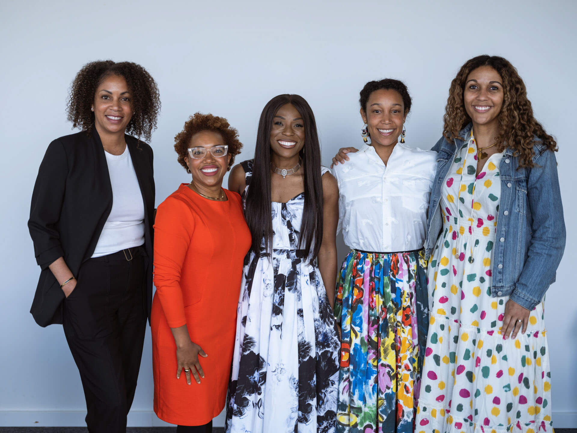 A group of five women posing together, dressed in vibrant and stylish outfits, smiling at the camera.
