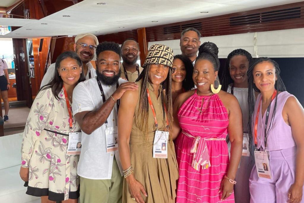 A group of adults gathers inside a yacht, smiling for the camera. They wear colorful summer outfits and conference badges, standing closely together in a bright interior space.