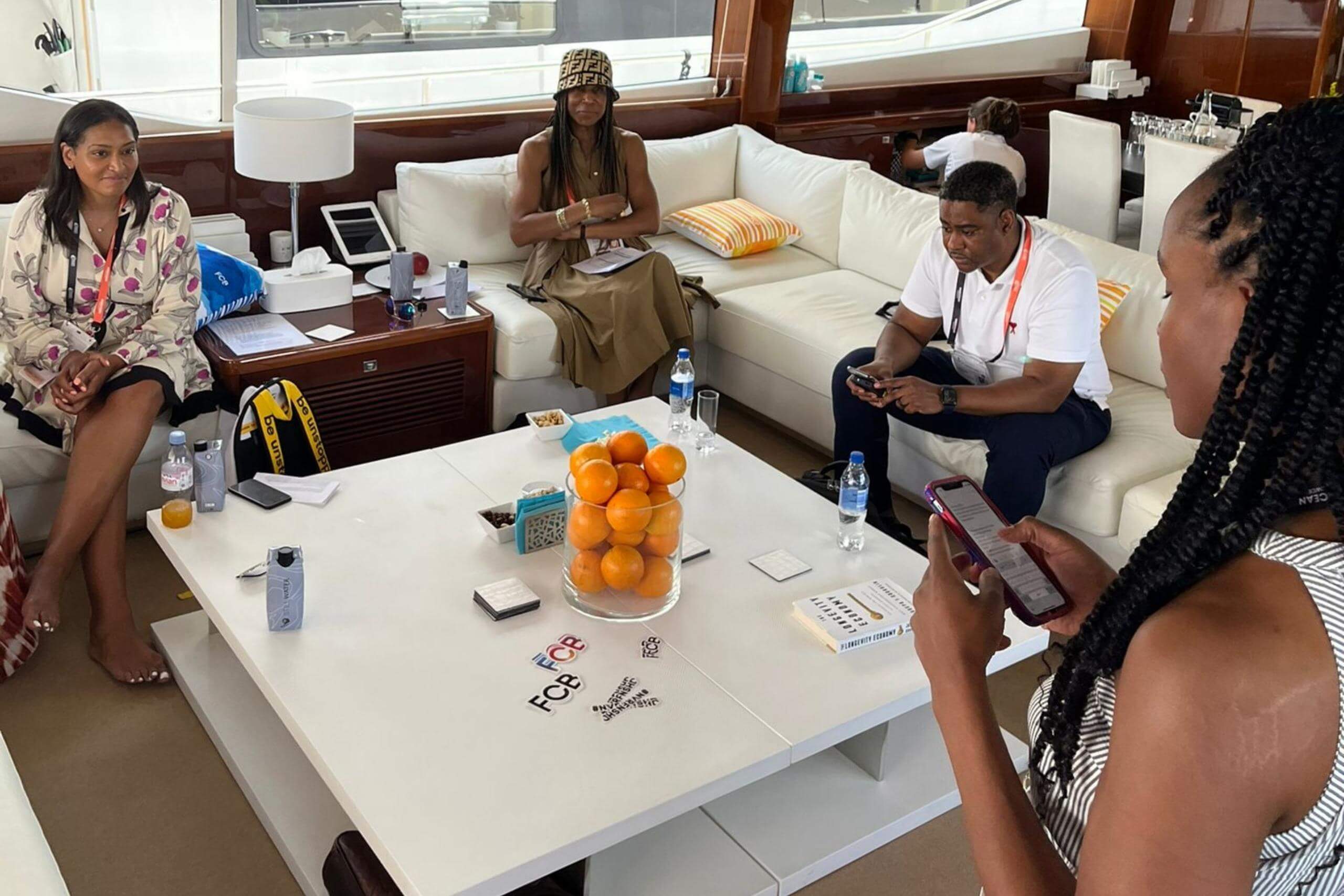 A small group seated on a yacht around a white table with snacks and documents. One participant is standing and reading from a phone, fostering a collaborative environment.