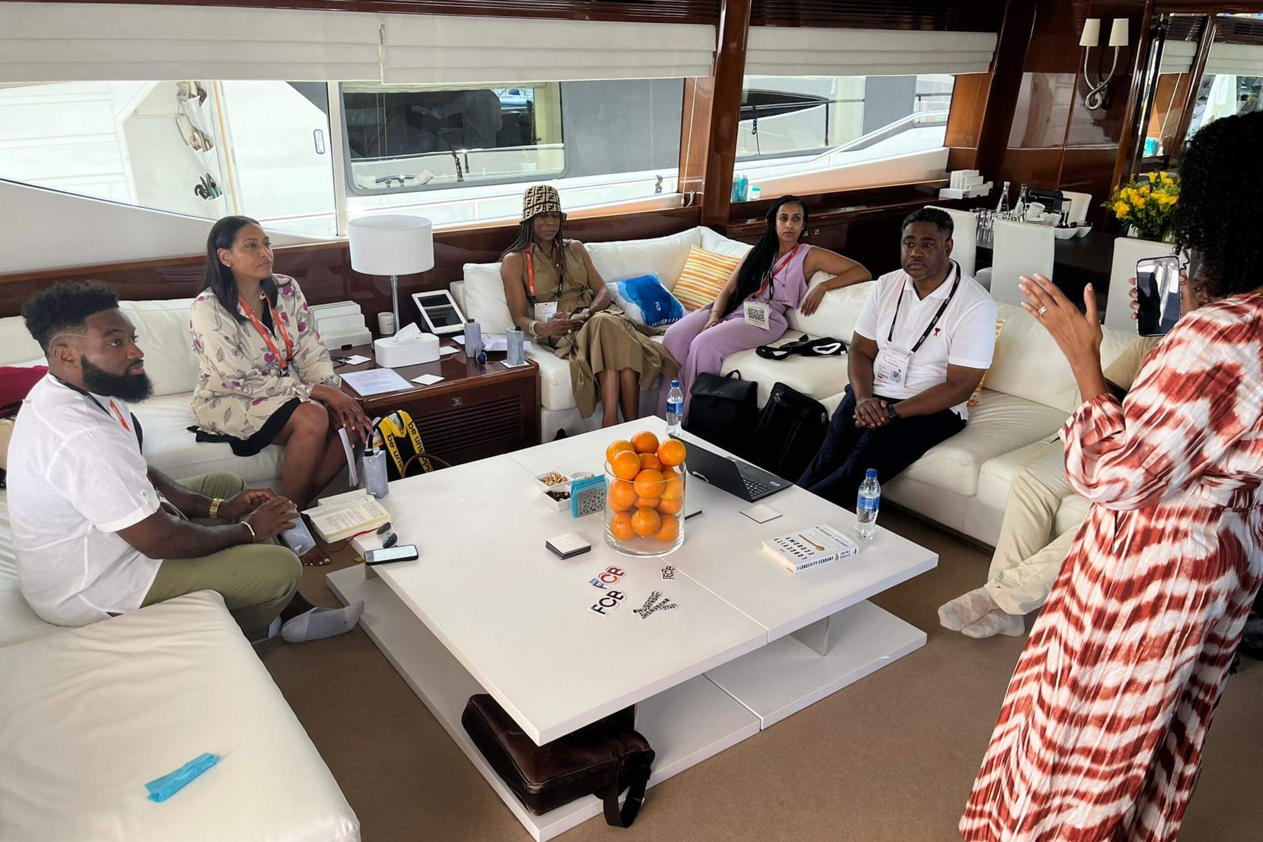 A broader view of the group sitting on a yacht, with a participant standing and speaking. A table in the foreground holds oranges, snacks, and documents, emphasizing an informal setting.
