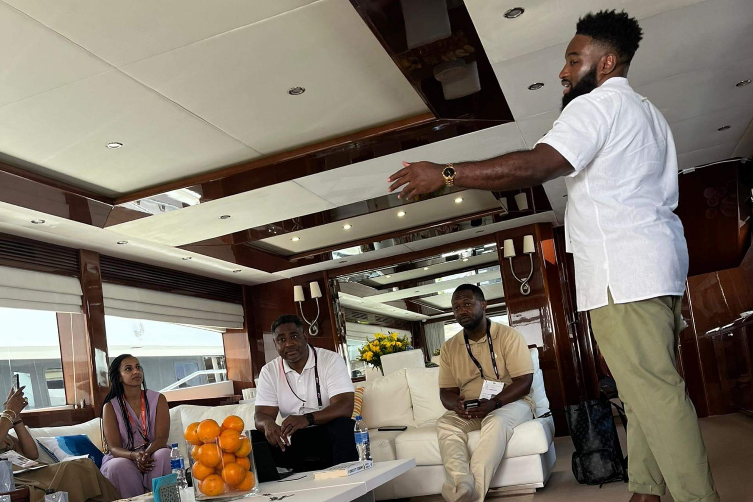 A speaker standing and gesturing during a discussion on a yacht. Other participants are seated, listening attentively in a relaxed setting with modern interiors.
