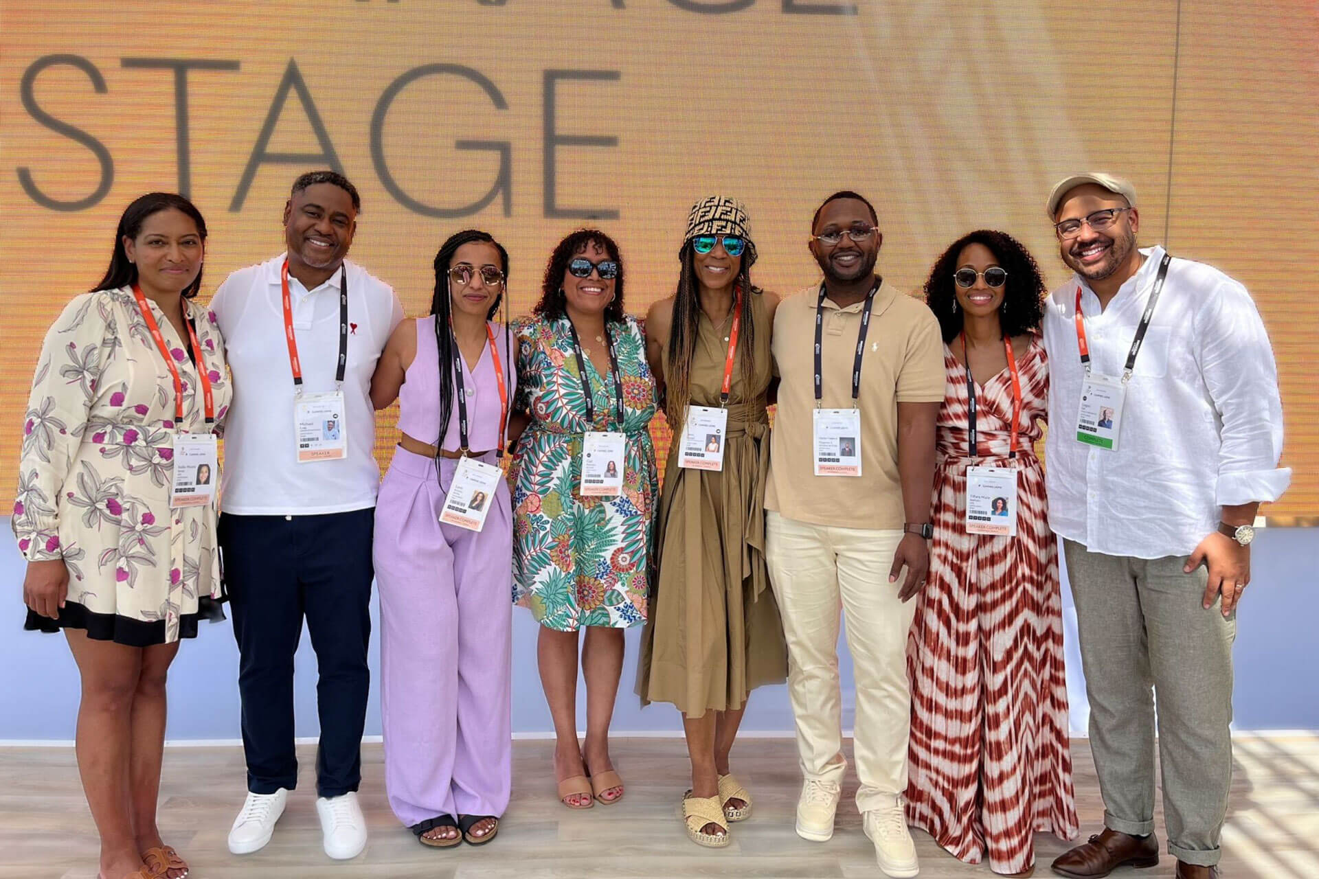 A group of eight people posing together at an event, all wearing badges and smiling for the camera. The backdrop features an event stage with a warm, professional setting.