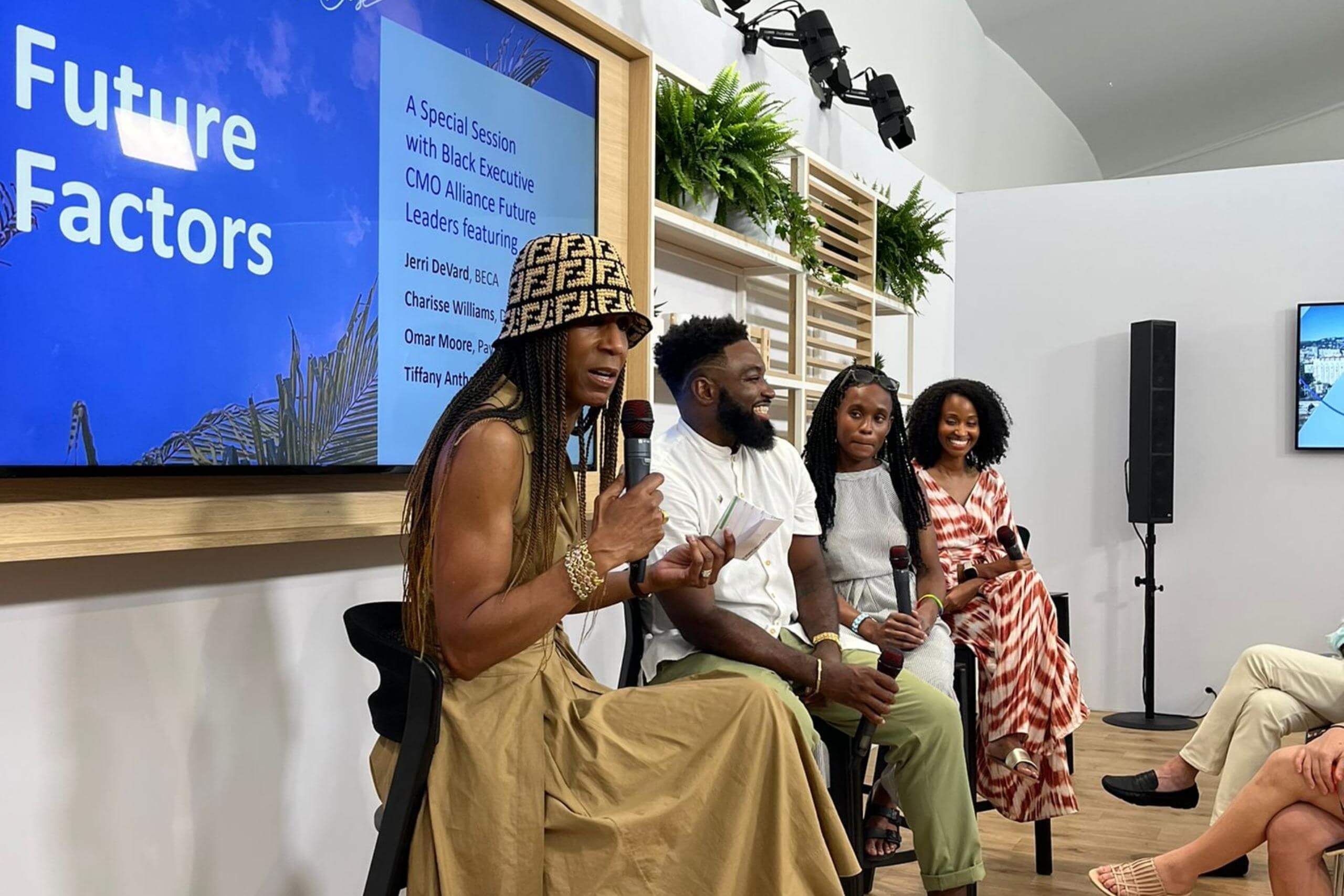 A panel discussion in progress, featuring four speakers seated on stools. One participant is speaking into a microphone, with a screen in the background displaying session details.
