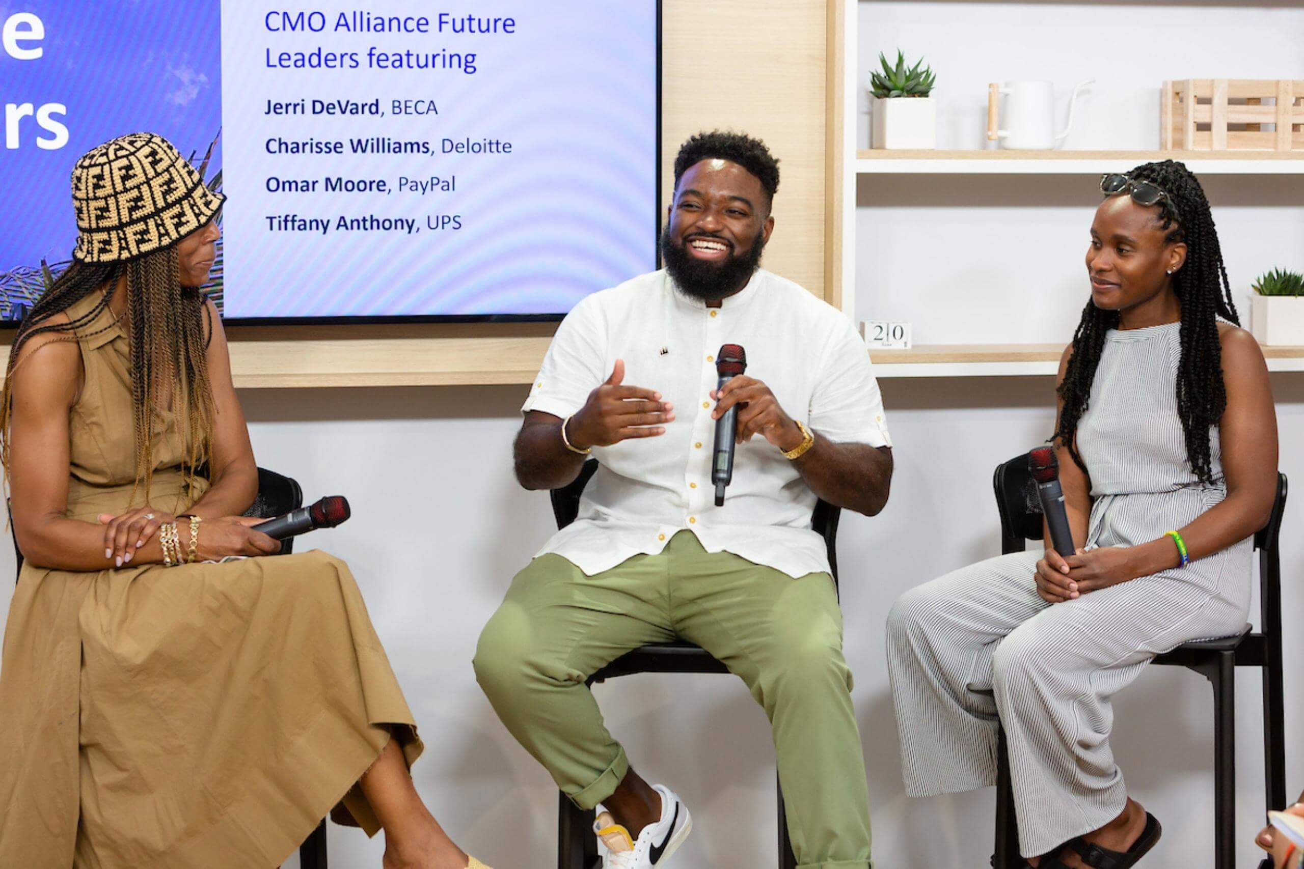 Three panelists sit on stools, engaging in discussion. One speaker gestures while holding a microphone, and the screen in the background lists the panel participants.