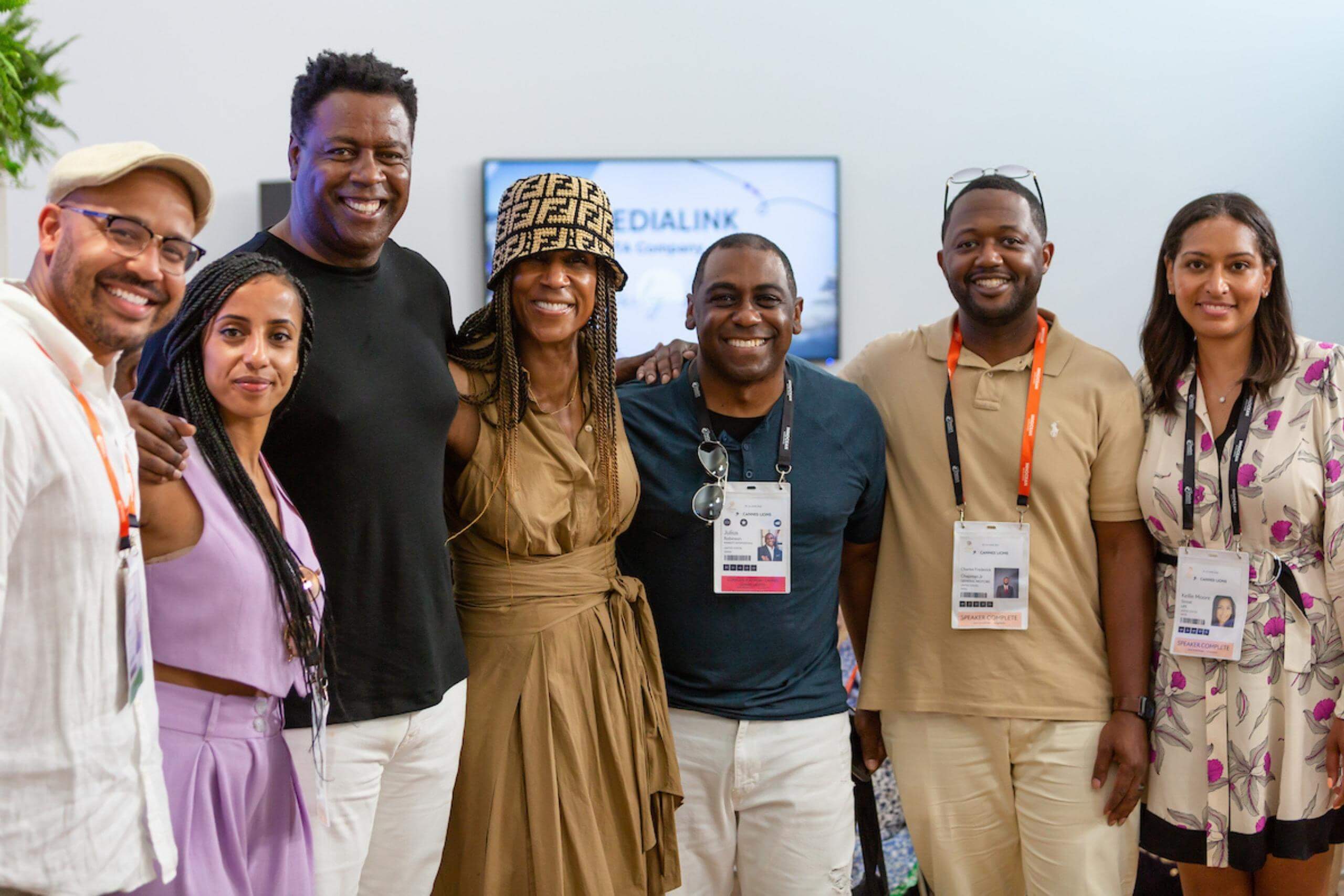 A group of seven people standing together indoors, smiling at the camera. They appear to be at a professional event, wearing badges and casual yet stylish attire.