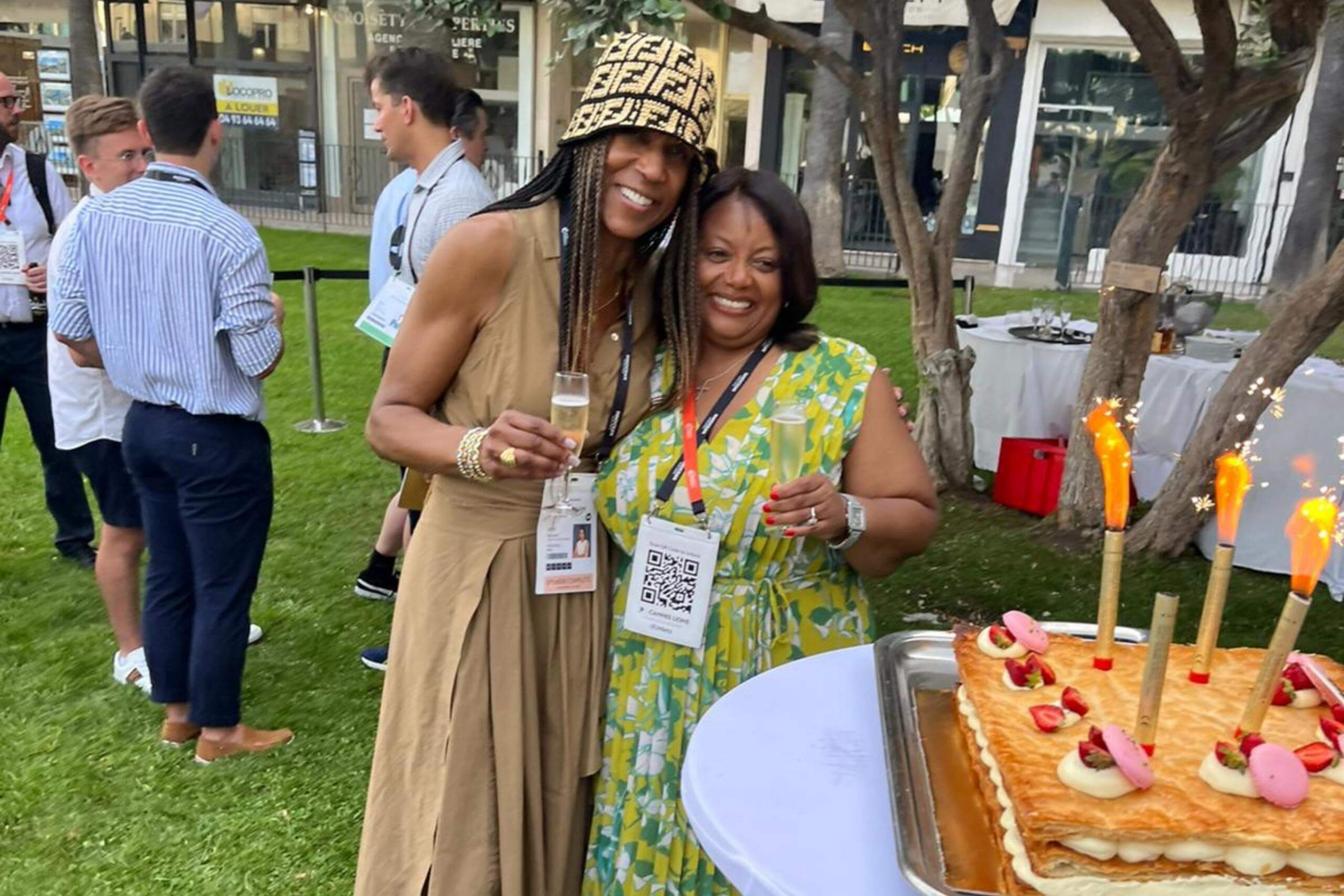 Two women outdoors at a celebratory event, holding champagne glasses and standing beside a cake adorned with macarons and lit sparklers. They are smiling and appear to be enjoying the occasion.
