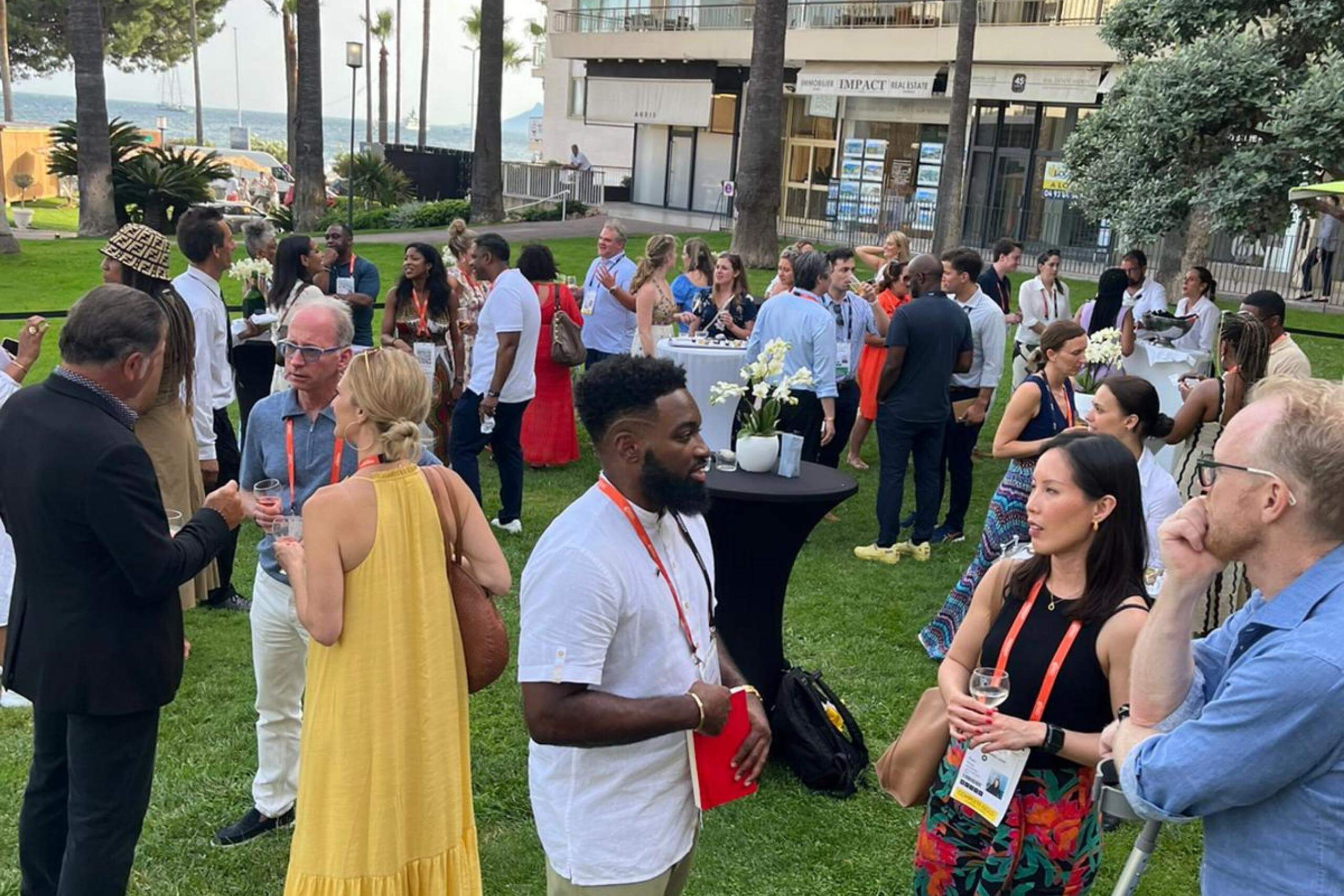 A gathering of people in a grassy outdoor area, mingling and conversing around high-top tables under a sunny sky.
