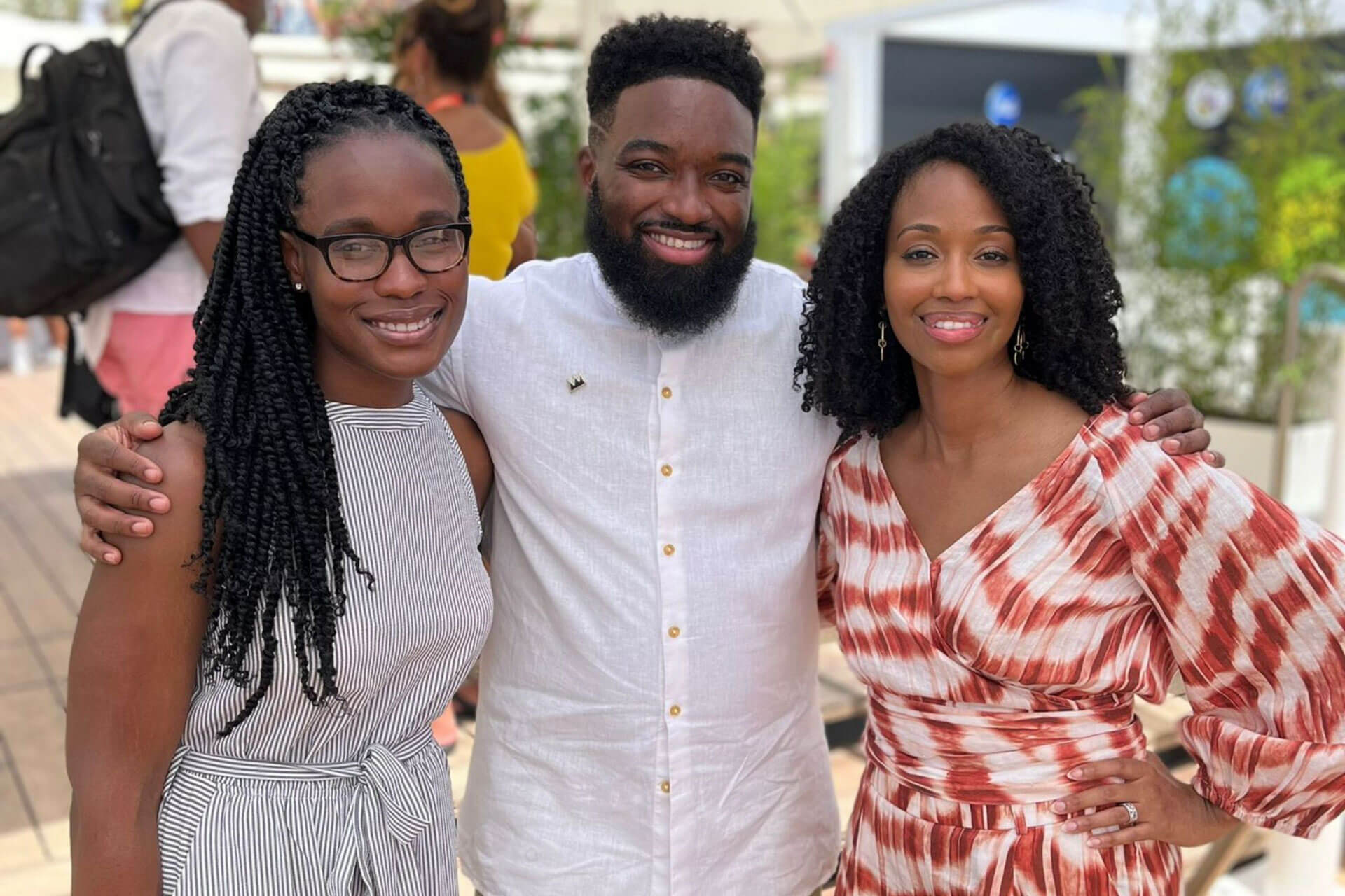 Three individuals standing close together, smiling warmly at the camera under a bright, sunny setting.