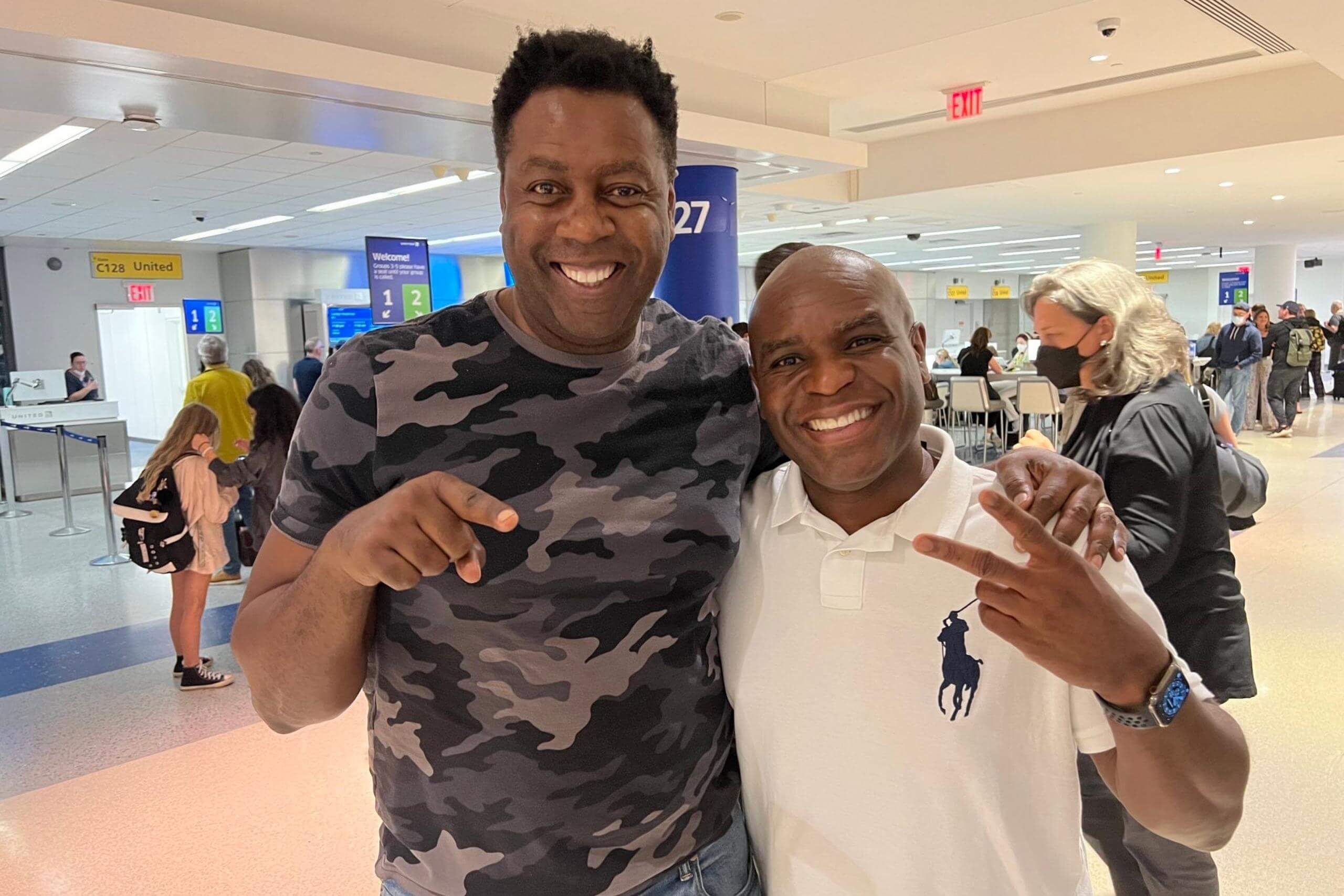 Two men smiling and posing together at an airport, one in a camouflage shirt and the other in a white polo.