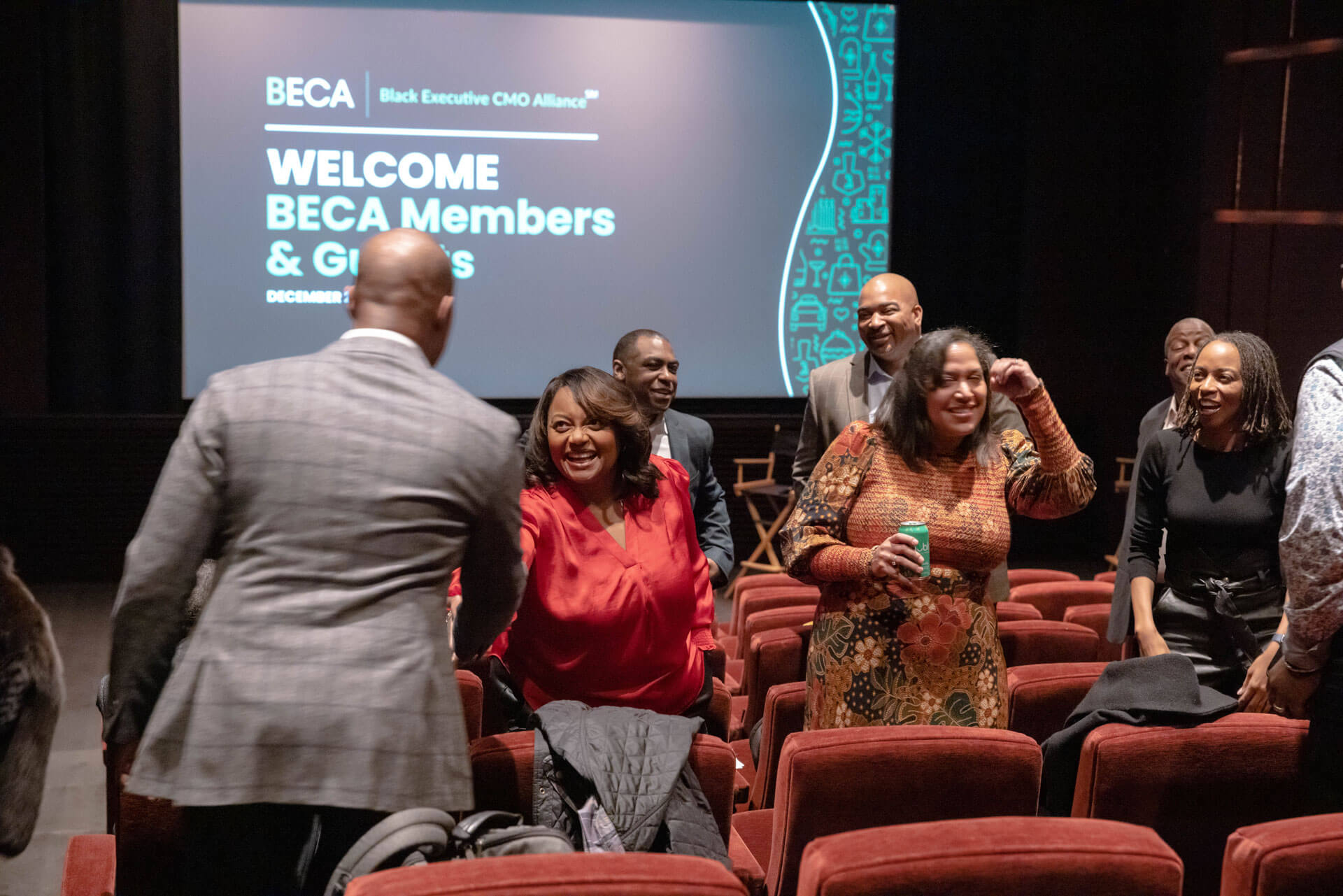 Event attendees mingling and greeting each other warmly in an auditorium with "BECA Members & Guests" projected in the background.