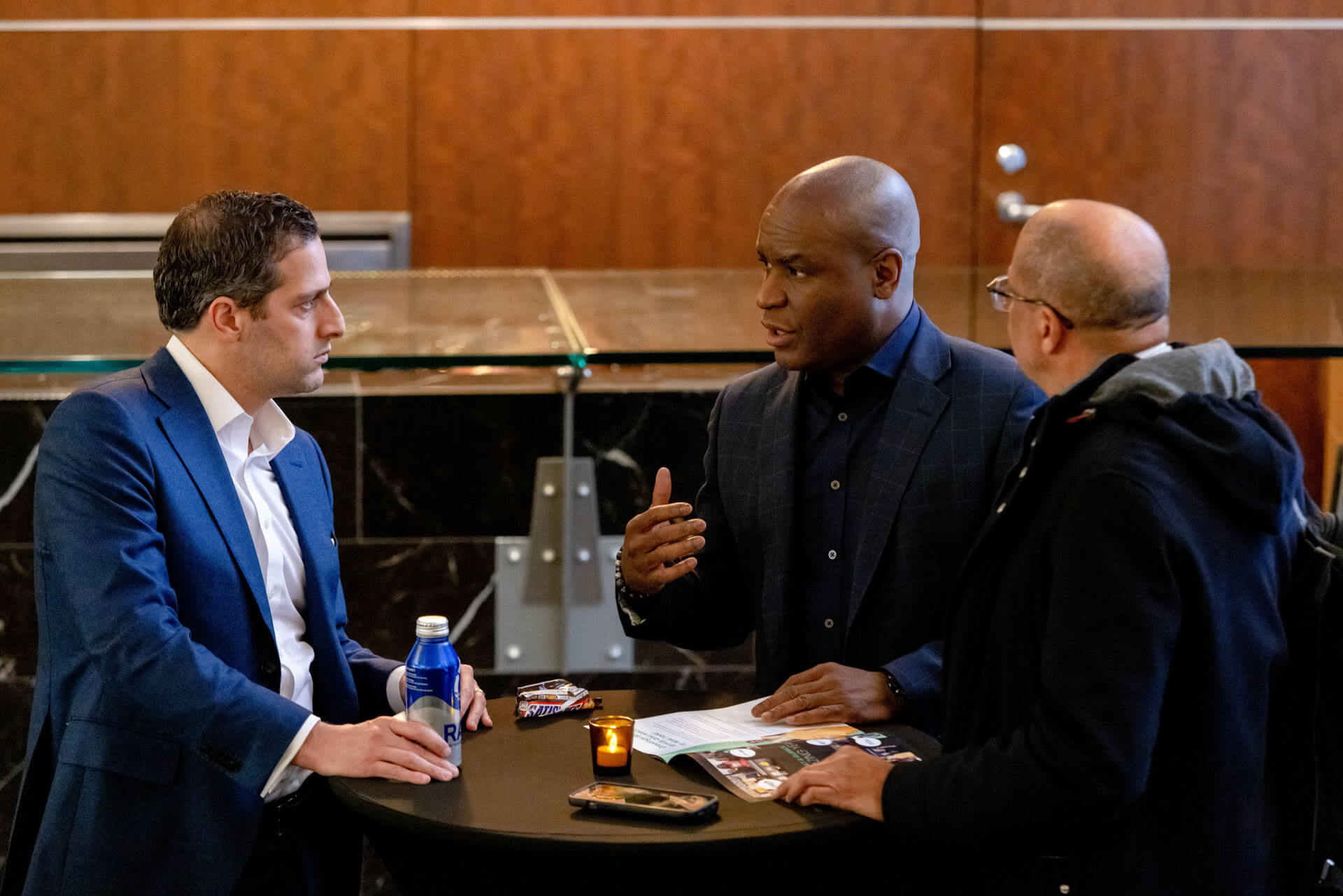 Three men in professional attire having an in-depth conversation around a high-top table in a modern setting.
