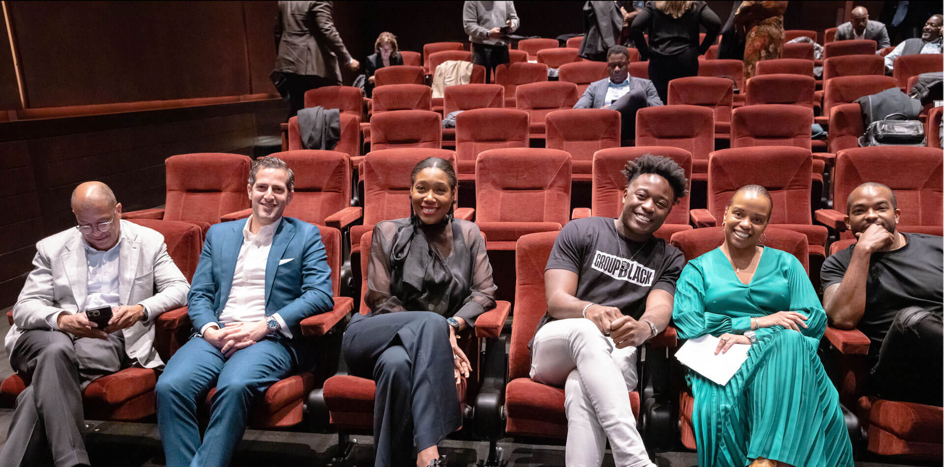 Six individuals sitting in a row of red theater seats, dressed in formal and semi-formal attire, with others visible in the background.