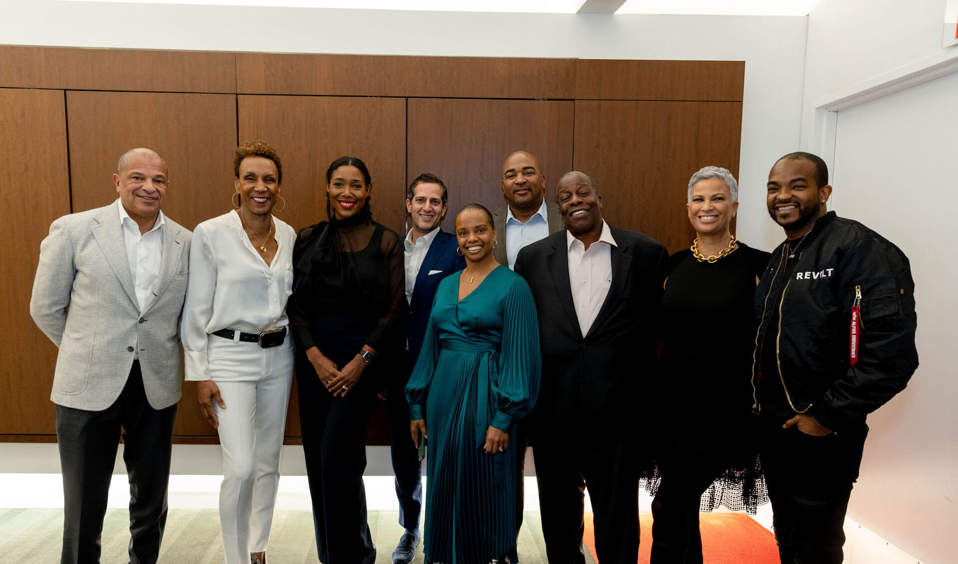 Nine individuals standing together, smiling, in formal and semi-formal attire in front of a wooden wall, representing a diverse group of professionals.