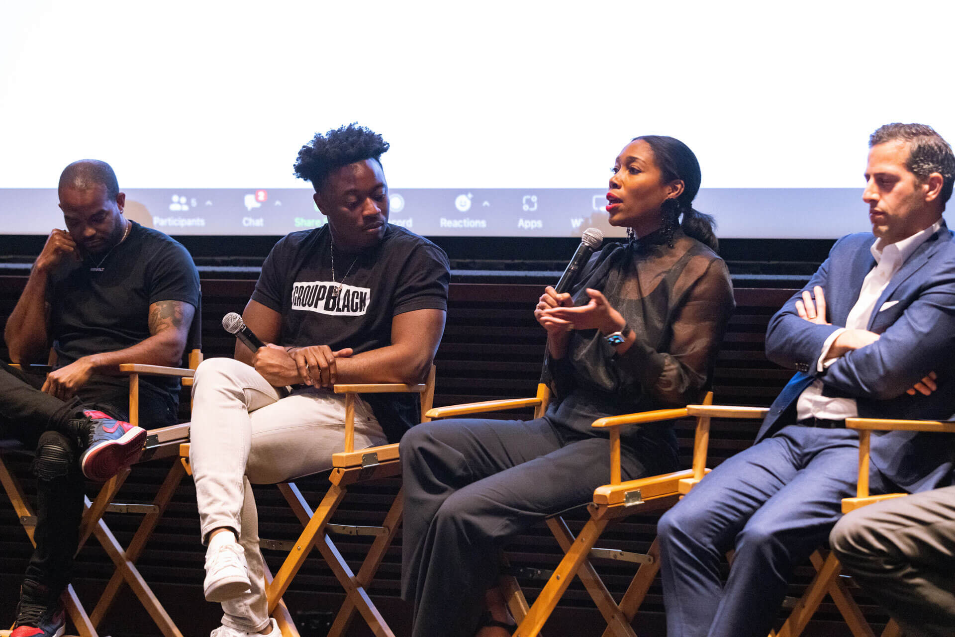 A panelist in a black outfit speaking into a microphone while others listen attentively, seated in director’s chairs with a screen in the background.