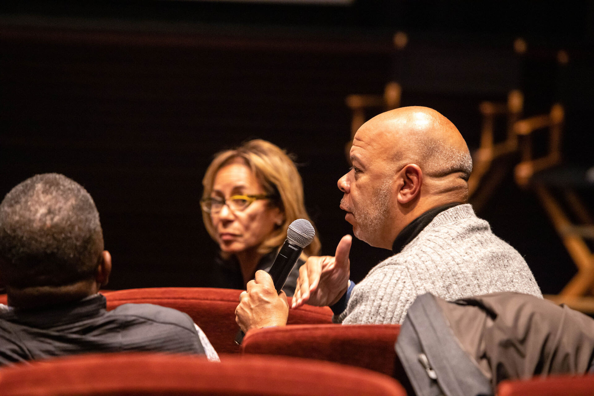A man sitting in a red theater chair holding a microphone, gesturing while speaking, with a woman in glasses attentively listening beside him.