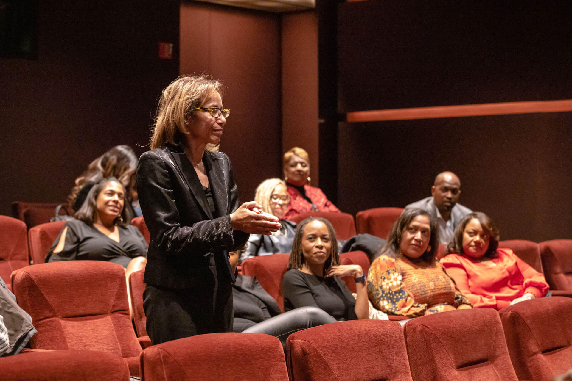 A woman in a black blazer standing in a theater, addressing the audience while others watch attentively.
