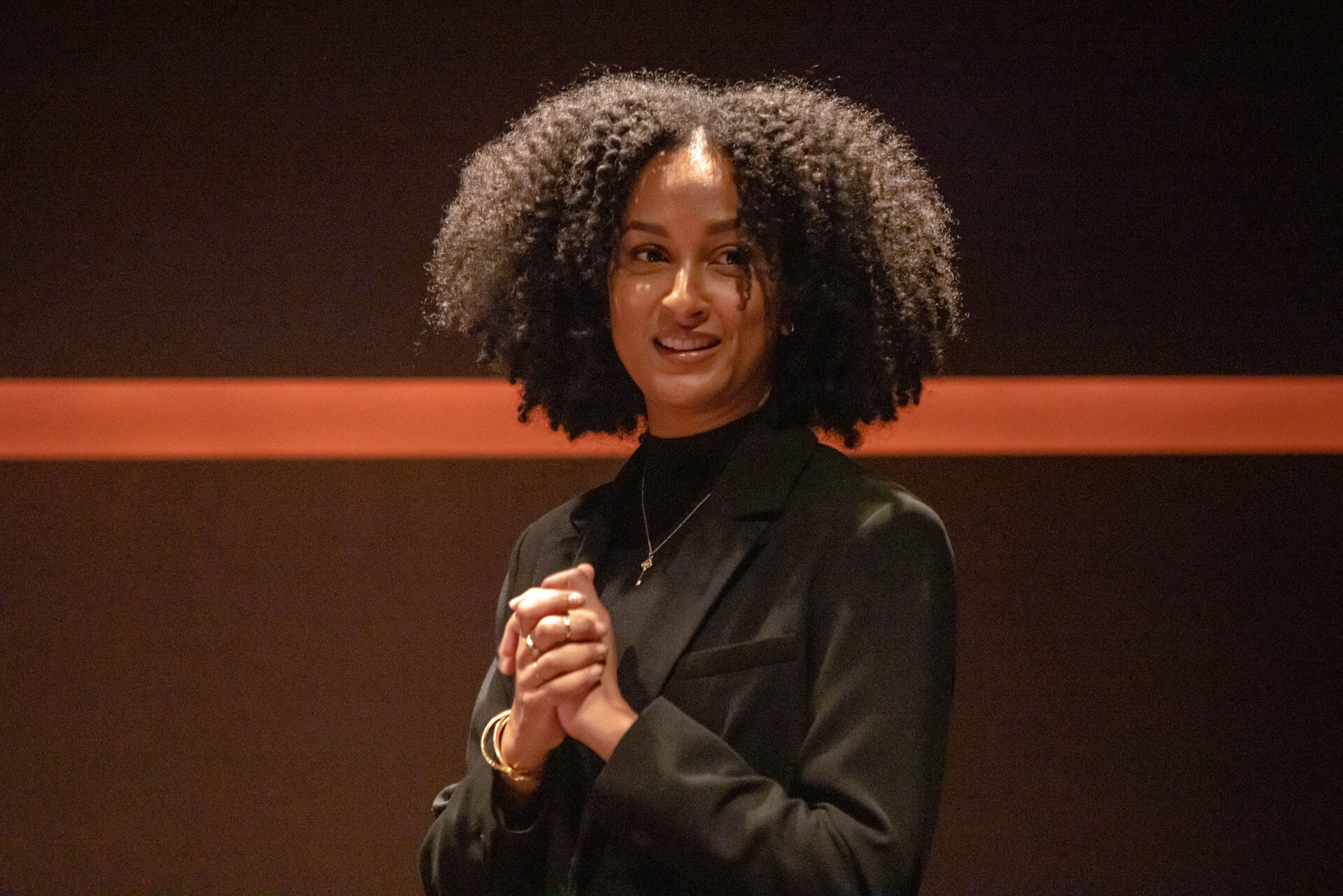 A young woman with curly hair standing, smiling gently, and addressing a group in a theater setting.