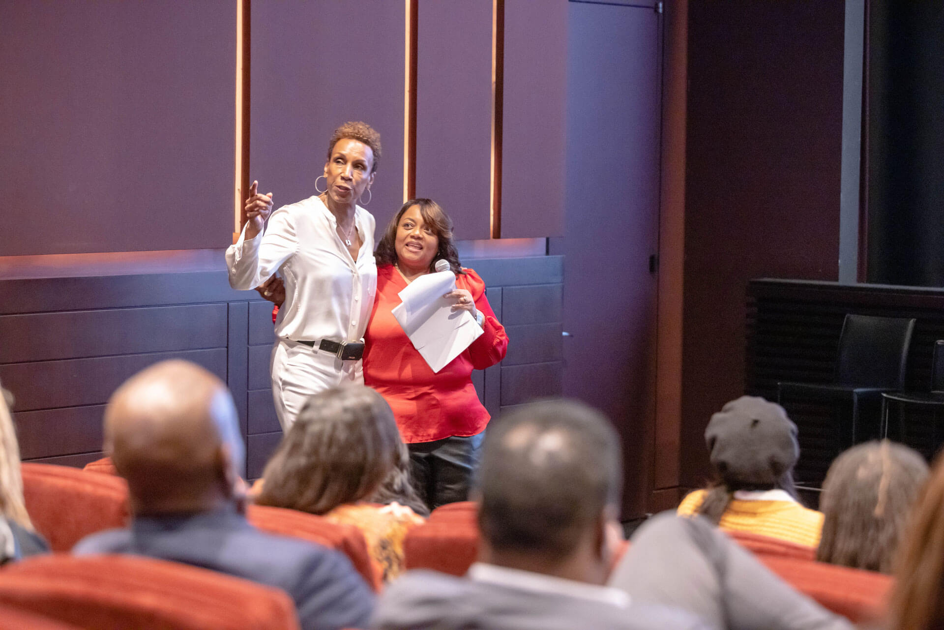 Two women standing at the front of a theater; one is speaking into a microphone while the other gestures toward the audience.