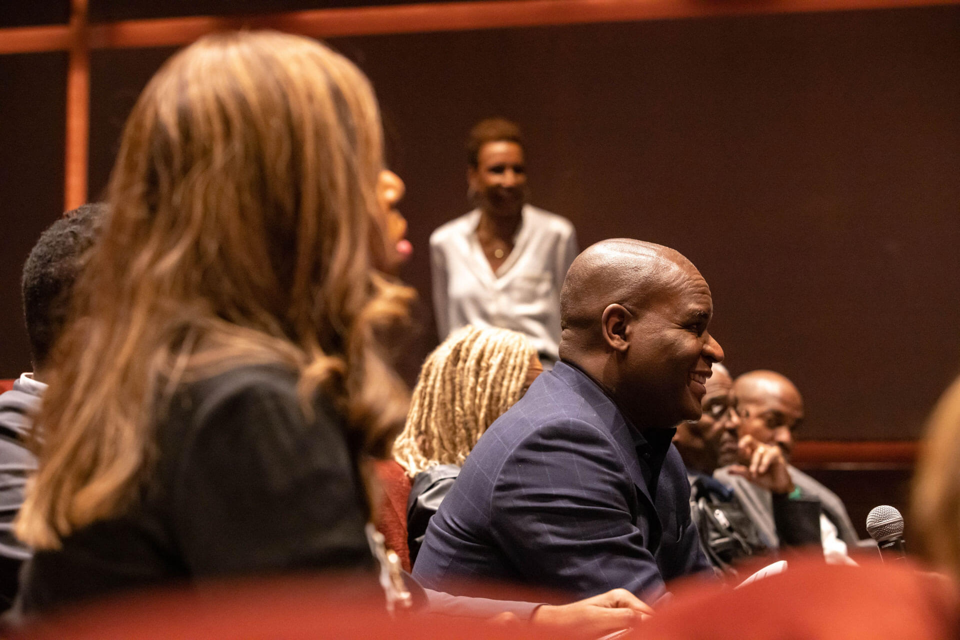 A man in a navy blazer sitting among other attendees in a theater, smiling during the event.
