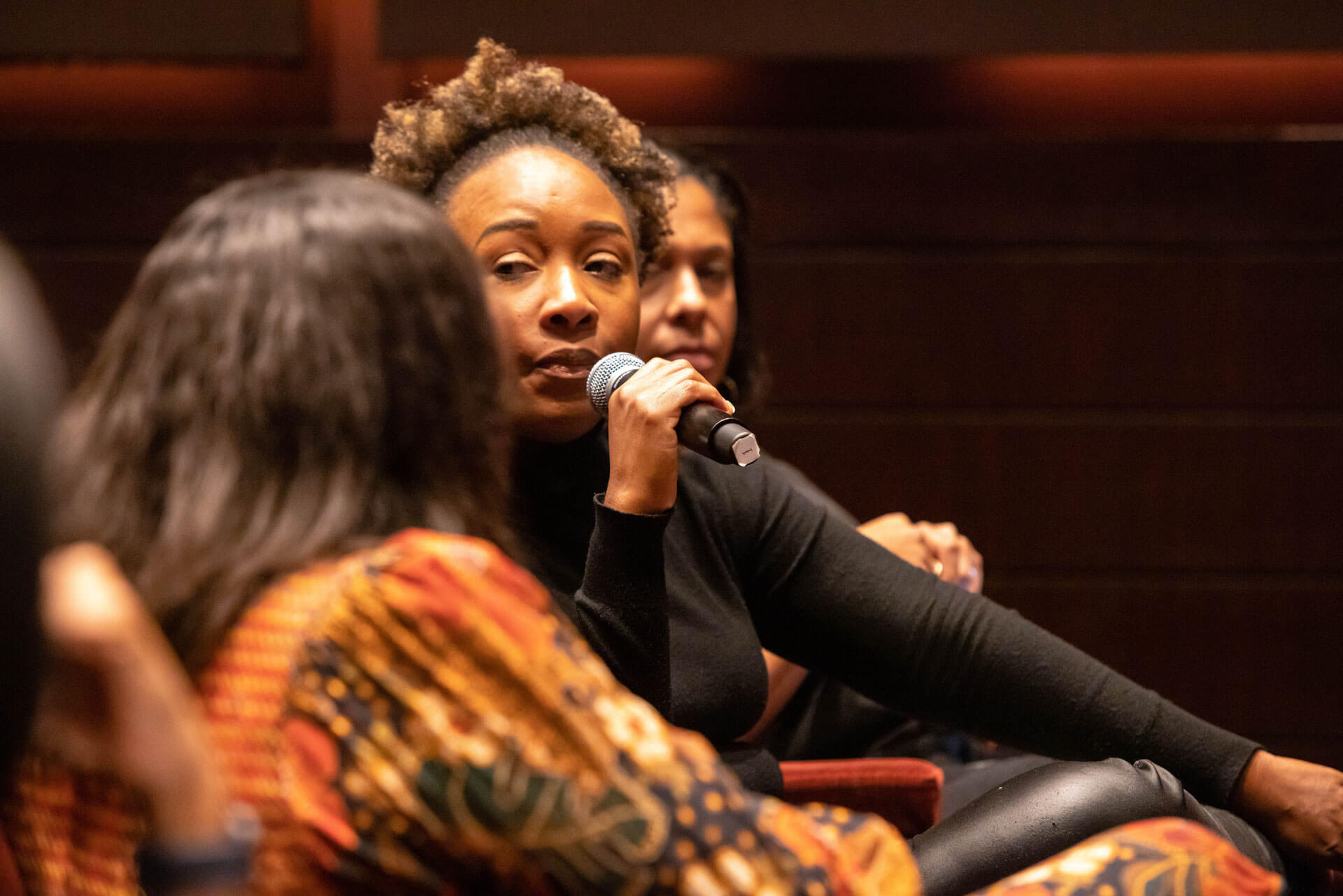A woman holding a microphone attentively participates in a discussion during an event, seated in a theater setting.