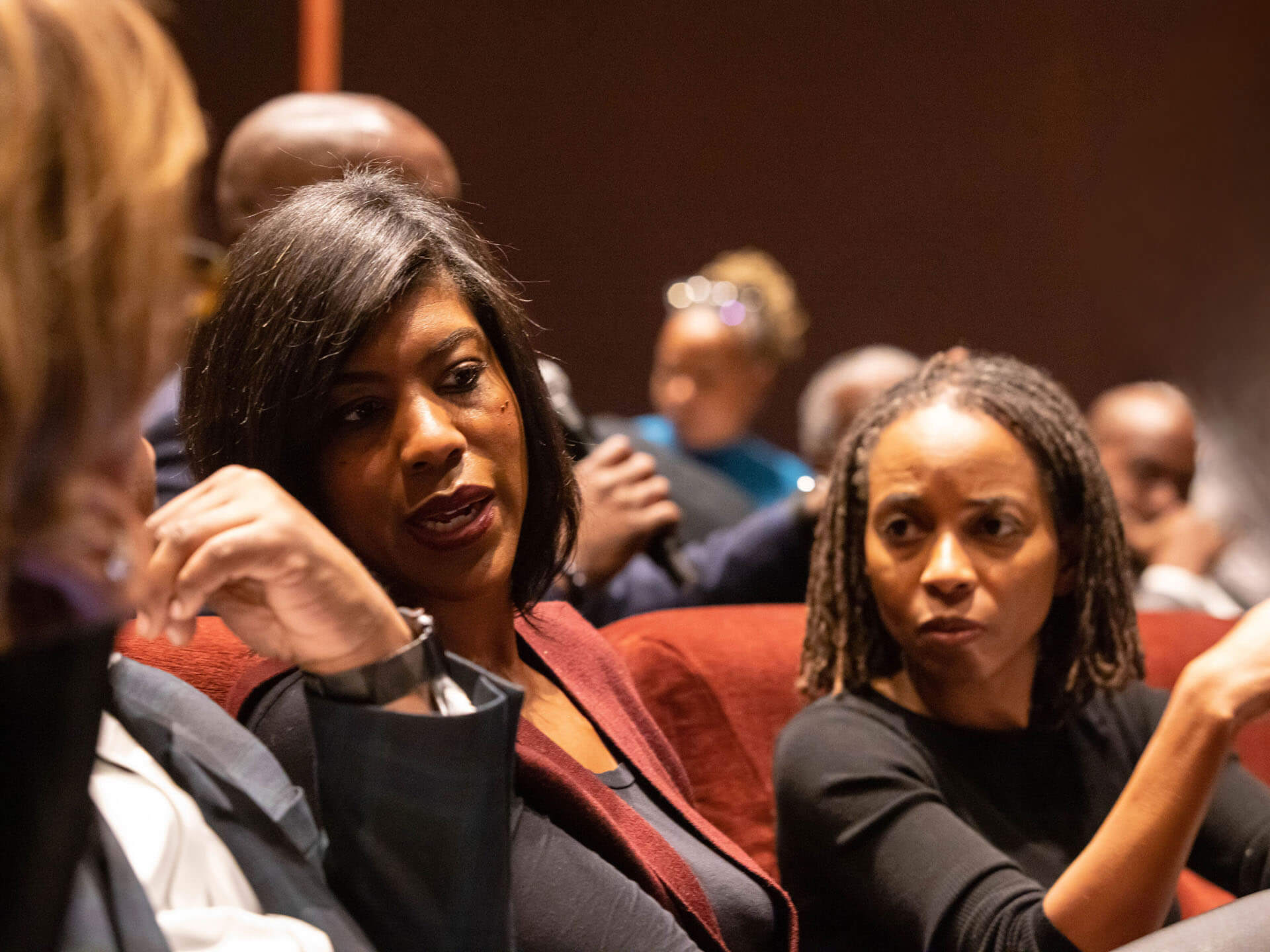 Two women engage in a deep conversation during a group discussion in a theater setting, with others seated nearby.