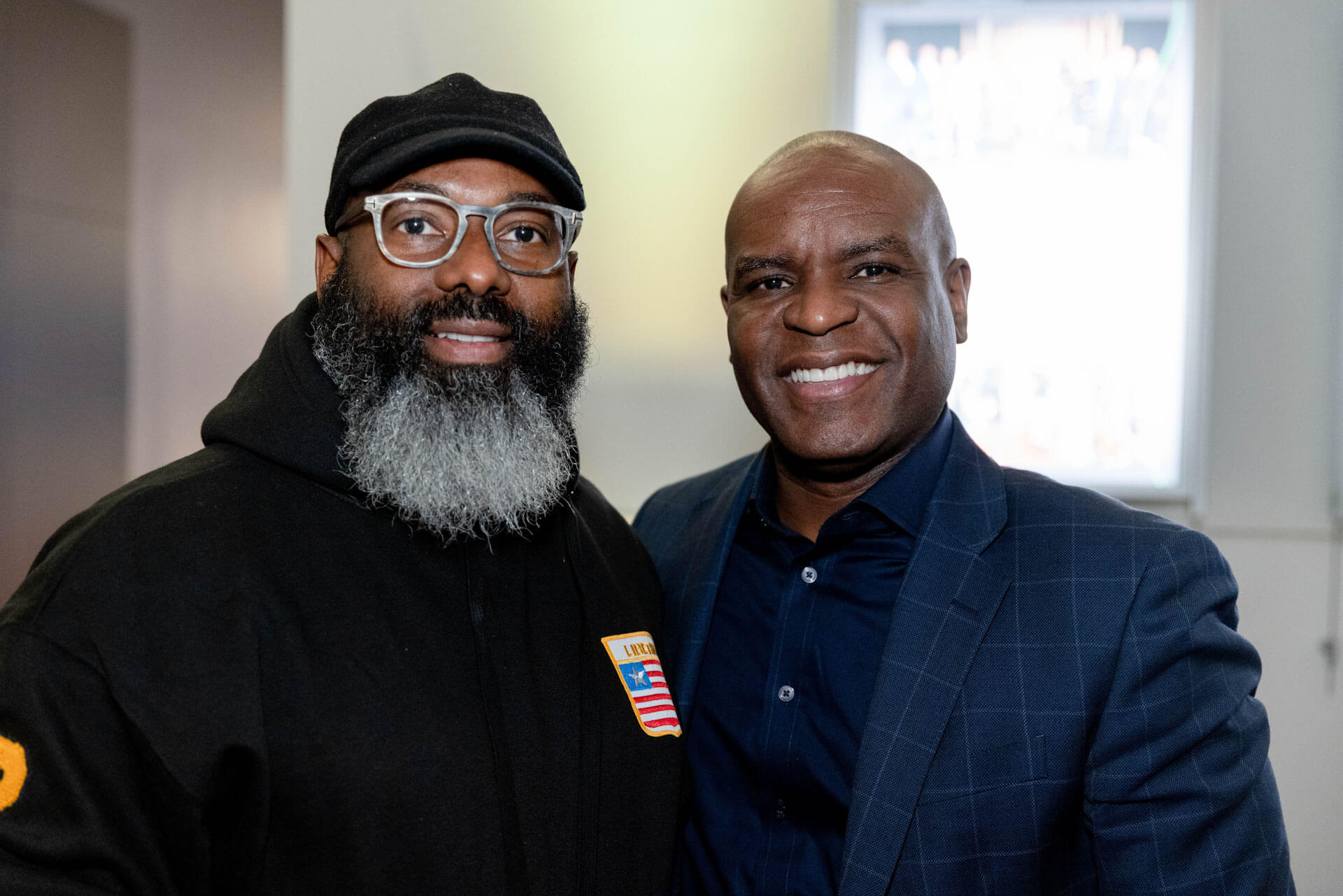 Two men pose together for a friendly photo, one wearing glasses and a hoodie, and the other dressed in a suit, smiling warmly.