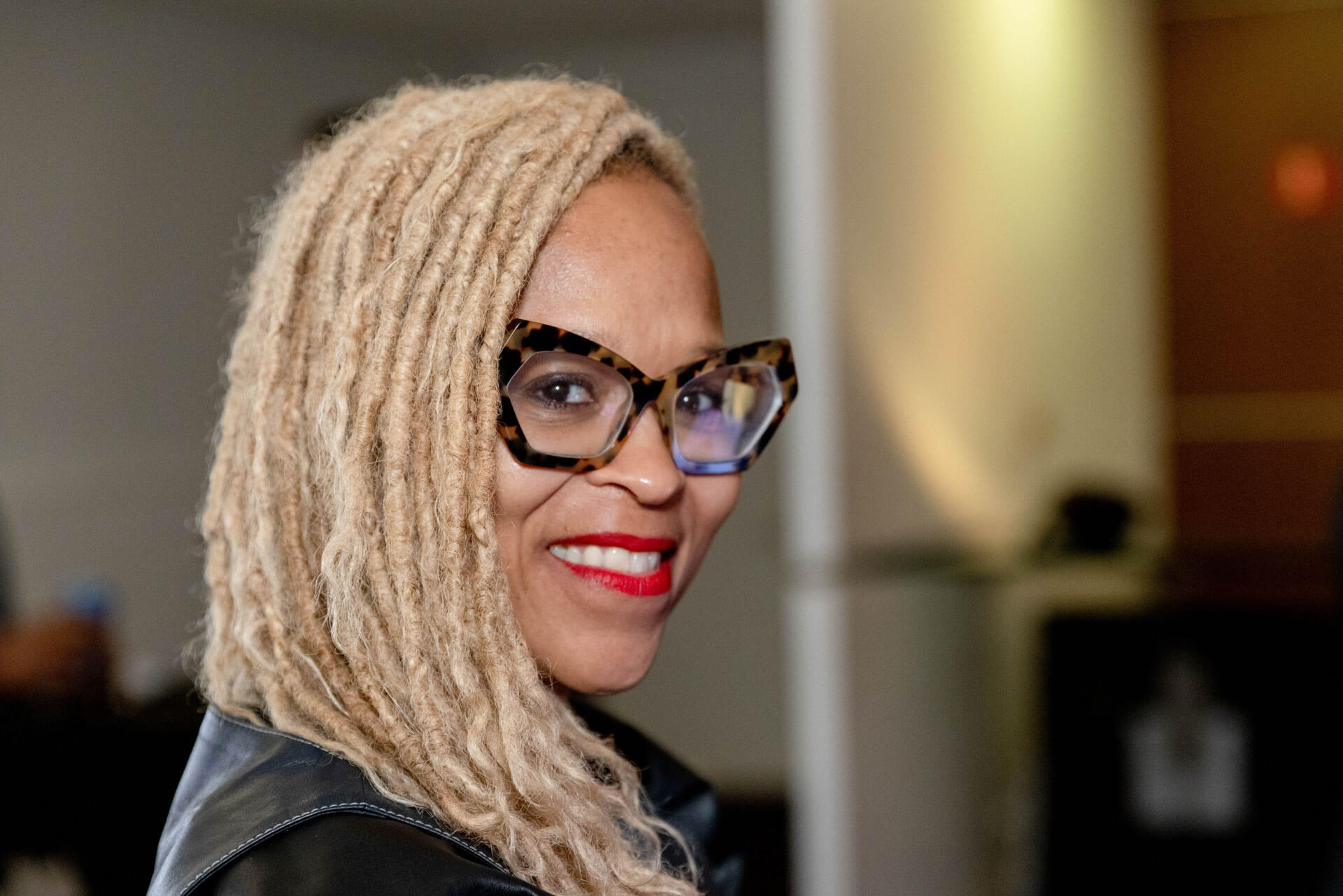 A close-up of a woman with blonde dreadlocks and stylish glasses, smiling confidently at the camera.