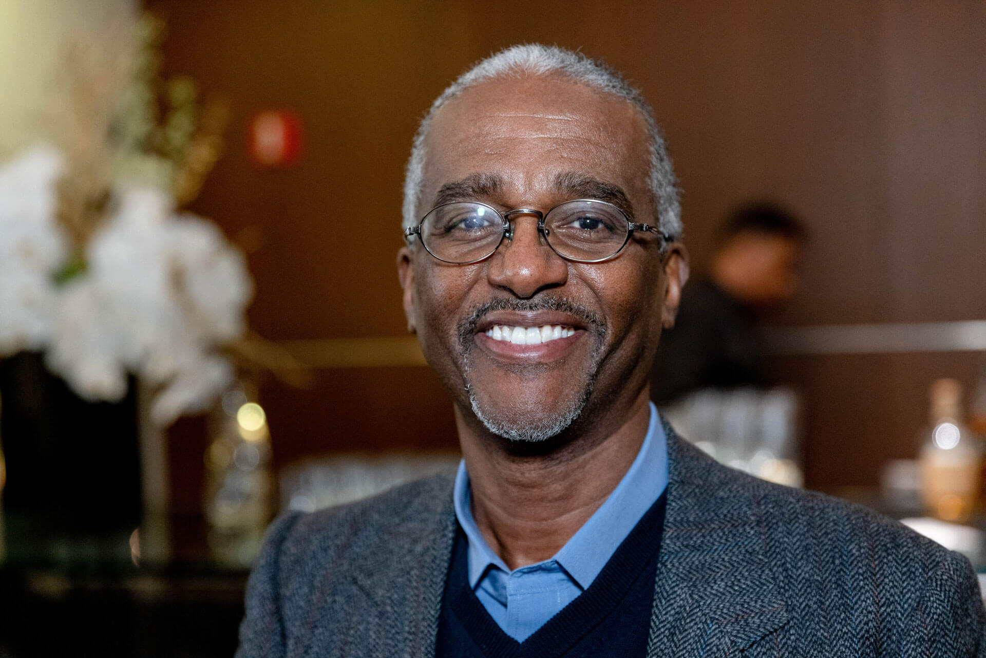 A man in glasses and a gray sweater smiles warmly in a professional portrait during an event.