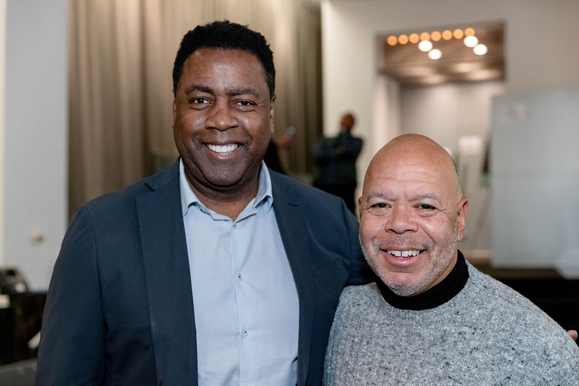 Two men pose side by side, smiling brightly, in a casual, indoor event environment.