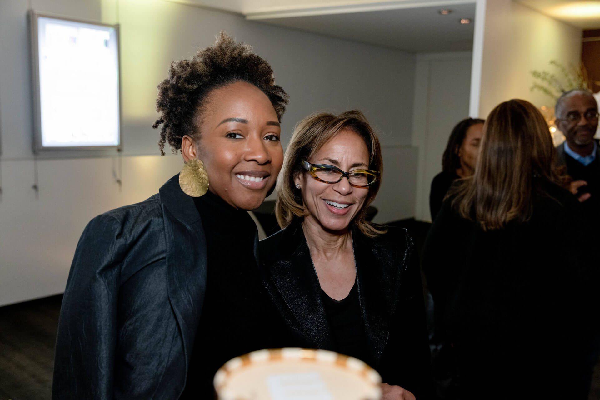 Two women share a moment, smiling at the camera during a networking or social gathering in a well-lit indoor setting.