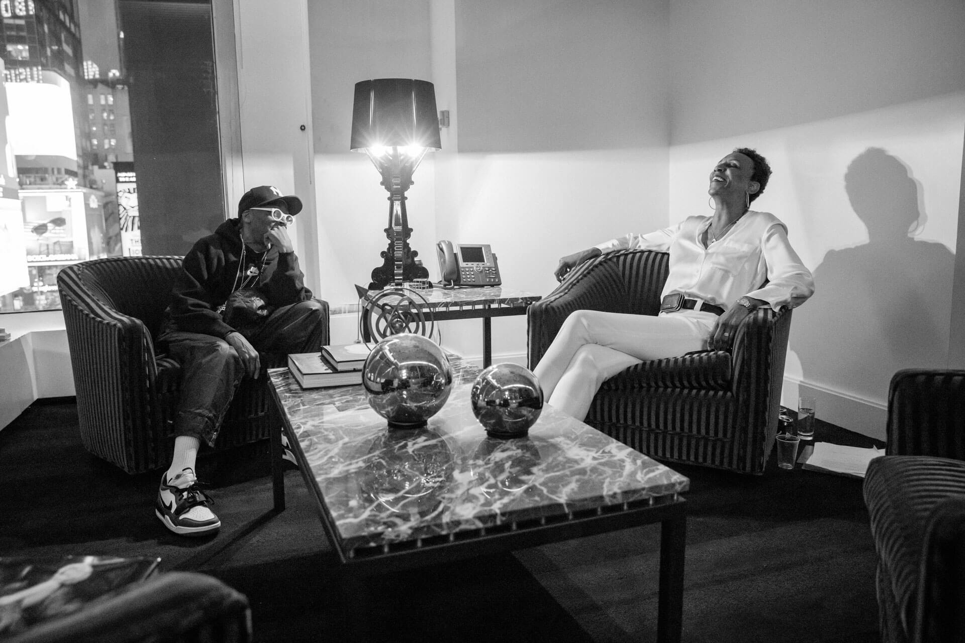 A black-and-white image capturing a casual, candid conversation between two individuals seated in a modern lounge area, one in a hoodie and the other in a stylish white outfit, sharing a moment of laughter.