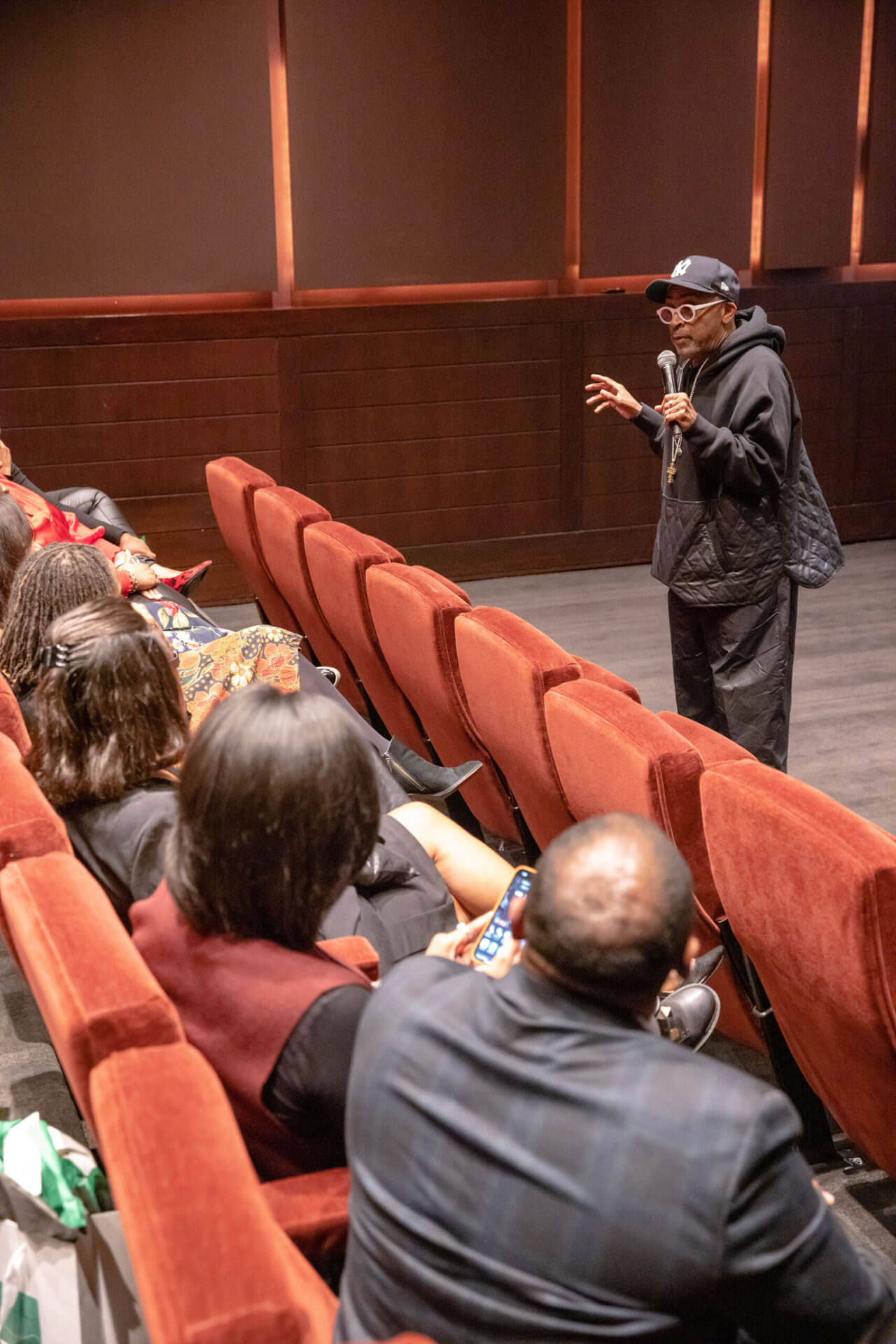 A speaker engages an audience in a theater, holding a microphone and gesturing animatedly during a presentation or discussion.